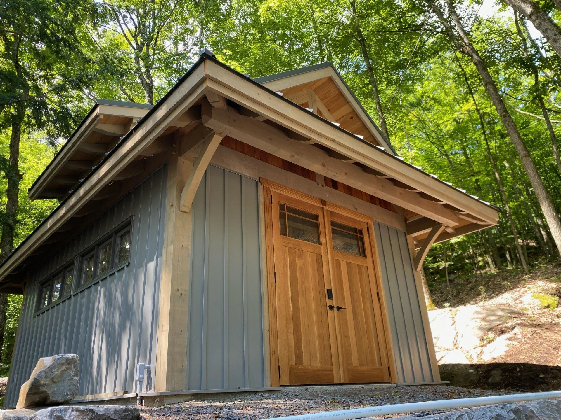 Small modern cabin with wooden doors and metal siding, situated in a wooded area with tall green trees.