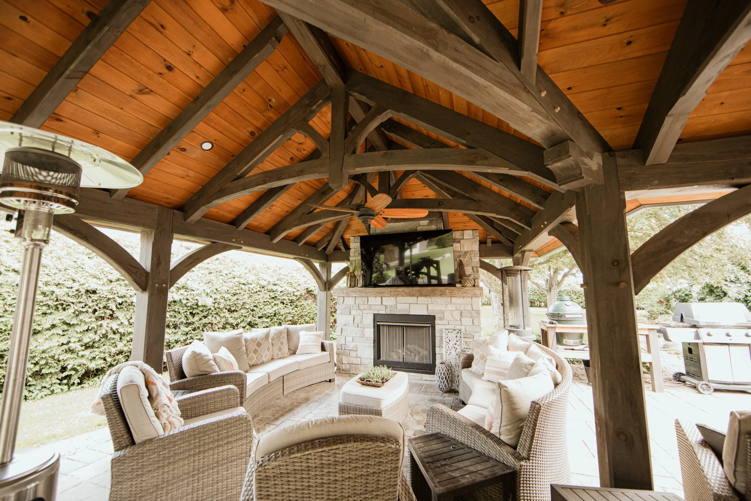 Outdoor living area under a wooden gazebo featuring a stone fireplace, a large TV, beige cushioned sofas, wicker chairs, and patio heaters, with lush greenery in the background.