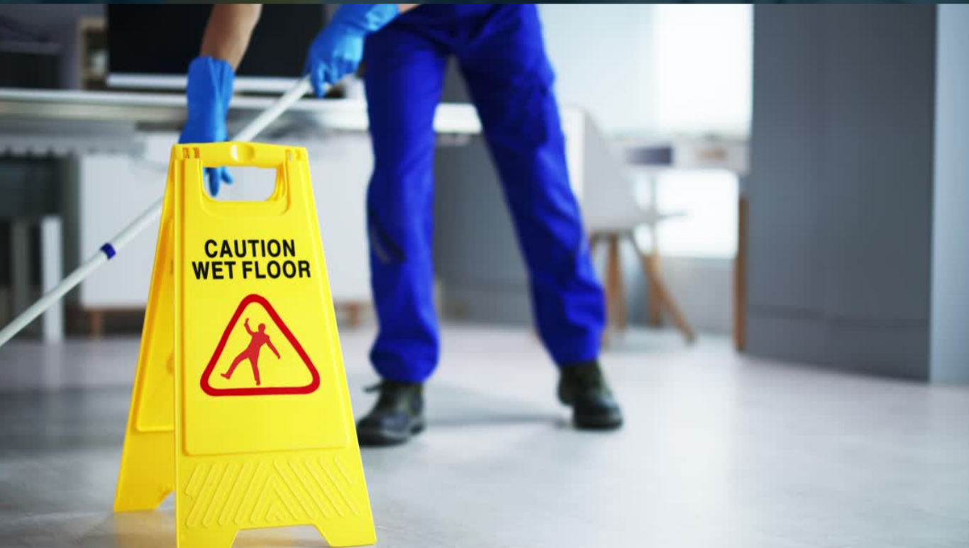 Person in blue uniform cleaning a wet floor with a mop, yellow caution sign that says "Caution Wet Floor" with a slip hazard icon.