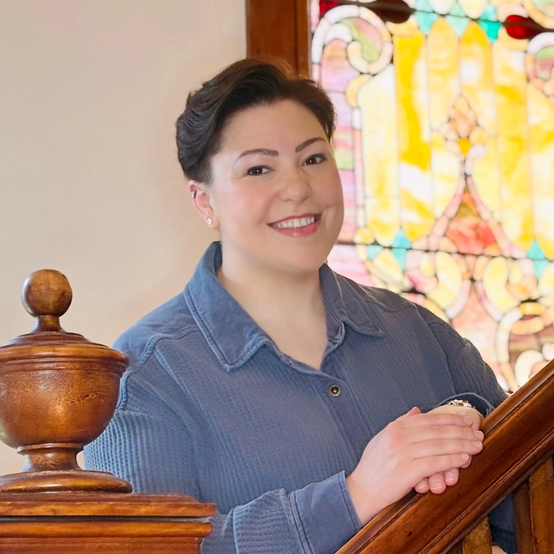 Abby stands on a wooden, Victorian staircase in front of a stained glass window. They have a dark, short haircut and are wearing a blue collared shirt.