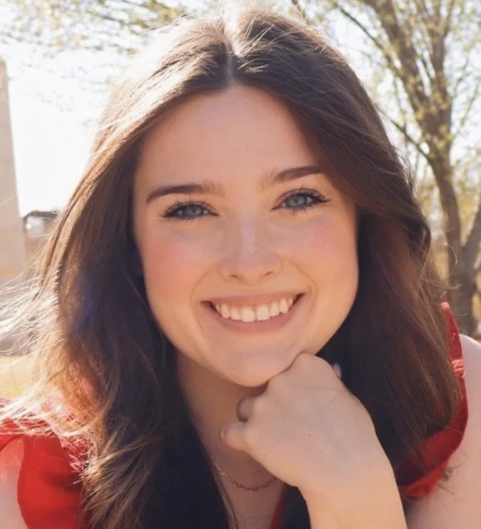 Smiling young woman with long brown hair, sitting outdoors in sunlight, wearing a red top.