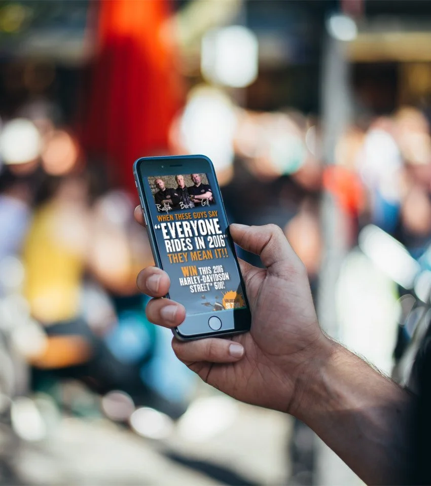 Person holding a smartphone displaying a promotional ad for a Harley-Davidson event in 2016.