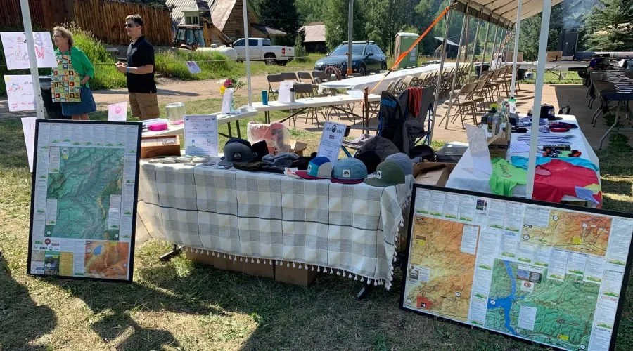 Outdoor booth at RTA fundraising event displaying maps, hats, and event materials, with people in the background near parked cars.