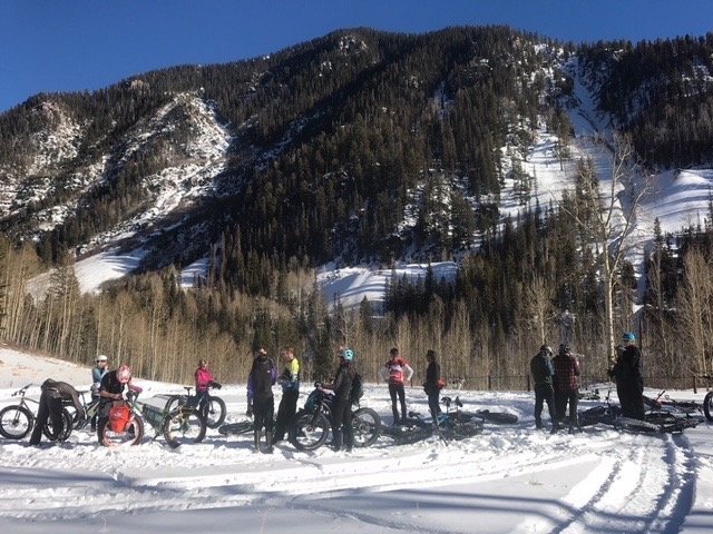 A group of fat bike riders and snow gear standing on snowy ground in front of a forested mountain landscape in Rico.