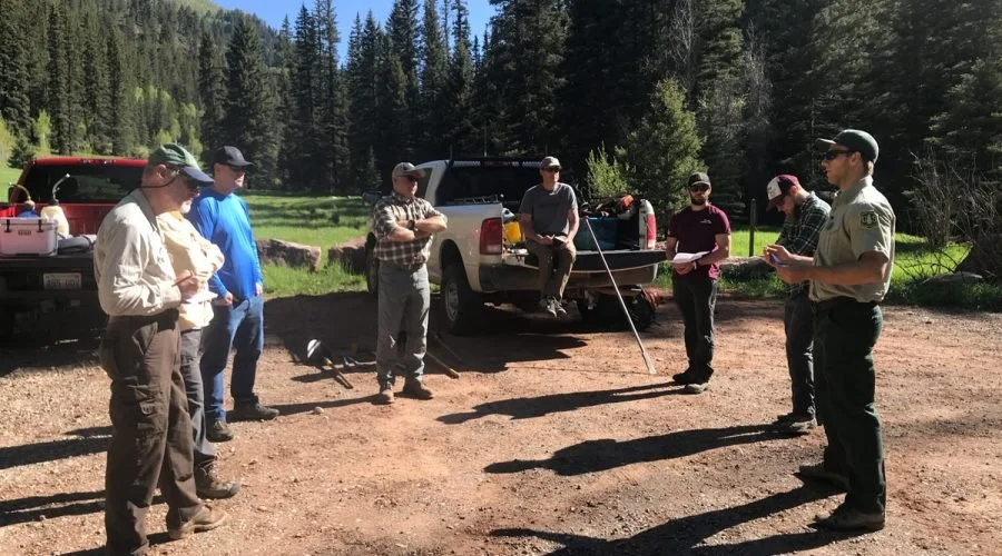 A group of volunteers, including a park ranger, gathered outdoors in a clearing surrounded by trees, with trucks in the background, preparing for a trail work day.