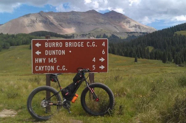 A mountain bike leaning against a sign for trails near Rico, Colorado in a grassy field with mountains in the background.