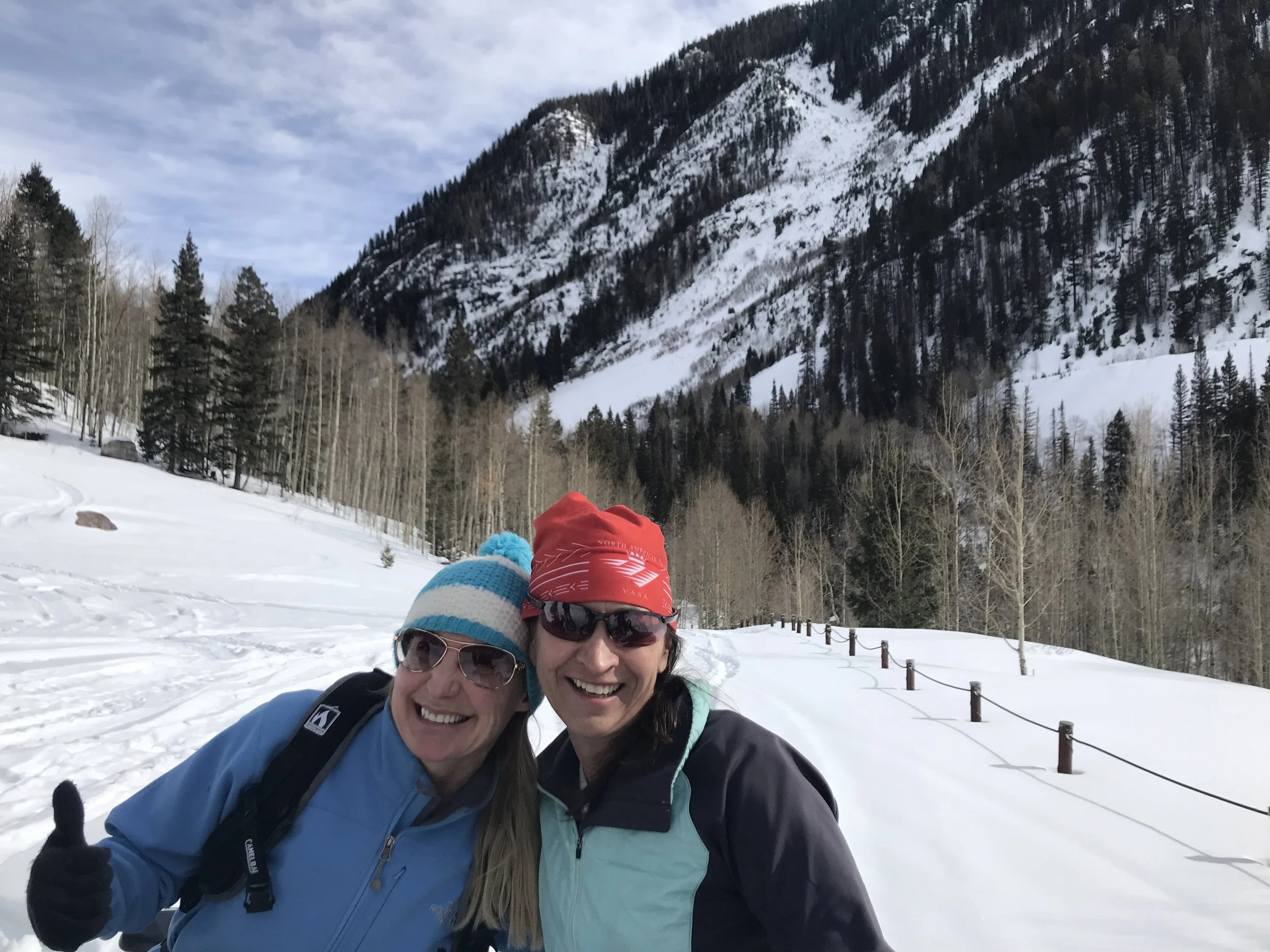 Two women smiling and dressed in winter gear, standing on a snow-covered trail in a mountainous forest landscape in Rico.