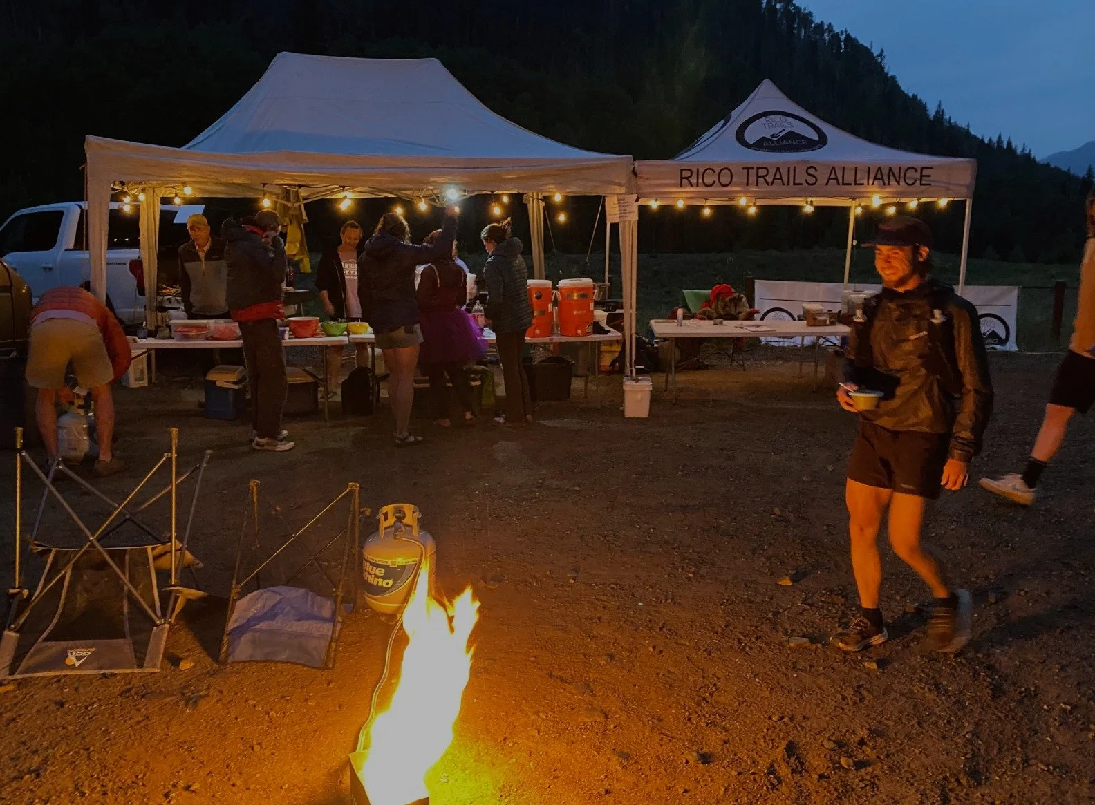 Tents labeled RICO TRAILS ALLIANCE, a small fire on the ground, and string lights illuminating the area, with mountains in the background at a fundraiser event.