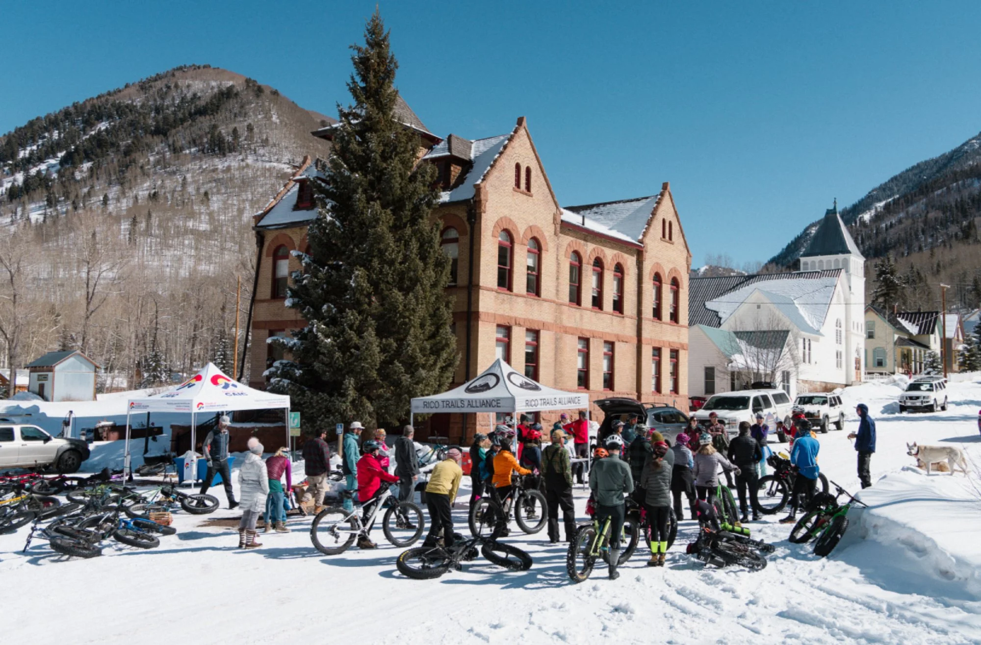 A snowy winter scene in a mountain town with a group of people gathered outside a historic brick building. Bicycles are parked, and tents from Rico Trails Alliance are set up. The background features the Rico courthouse and historic church buildings.