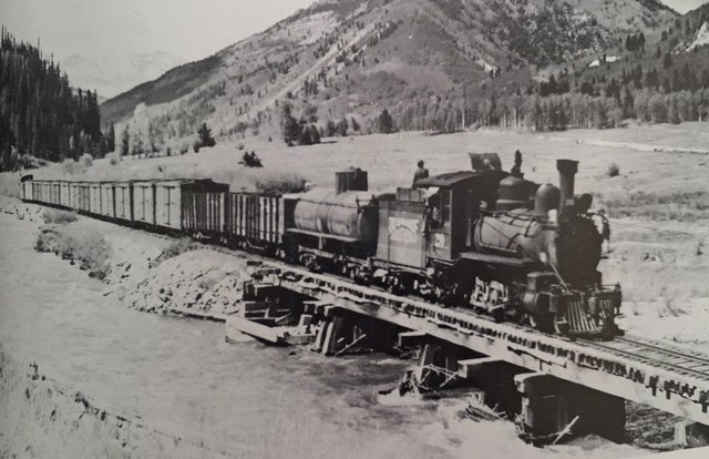 Black and white photo of a vintage steam locomotive train from Galloping Goose crossing a bridge over a river in Rico, Colorado.