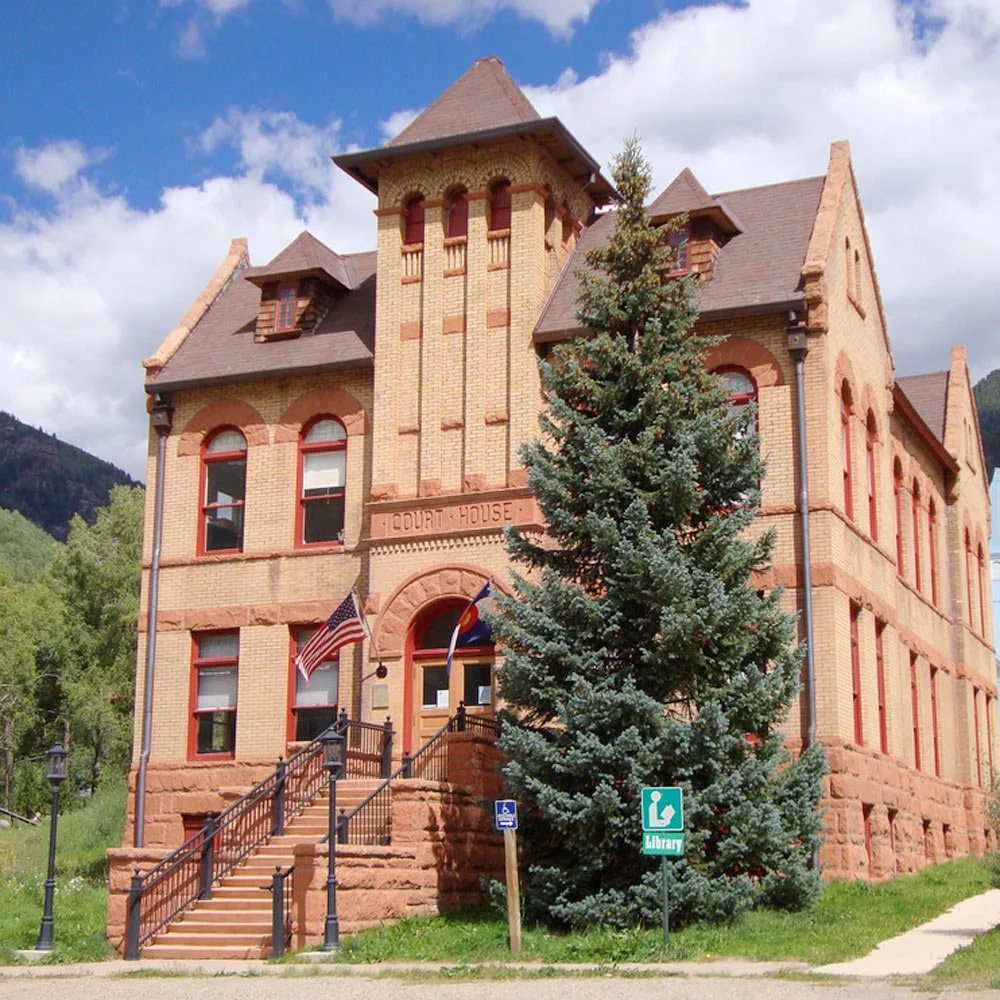Rico's historic brick courthouse building, set against a background of green mountains and partly cloudy sky.
