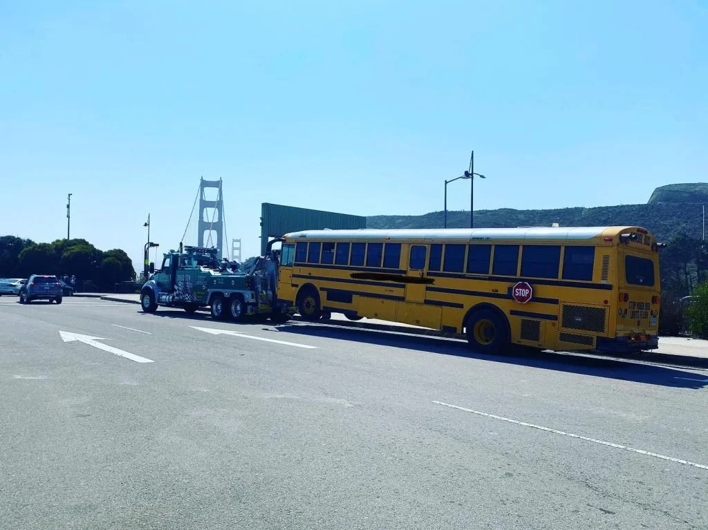 A yellow school bus with a stop sign on the side is being towed by a tow truck on a bridge with the Golden Gate Bridge in the background.