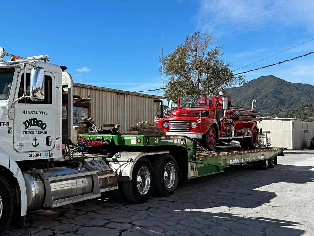 A red vintage fire truck loaded on a green flatbed tow truck outside with mountains in the background.