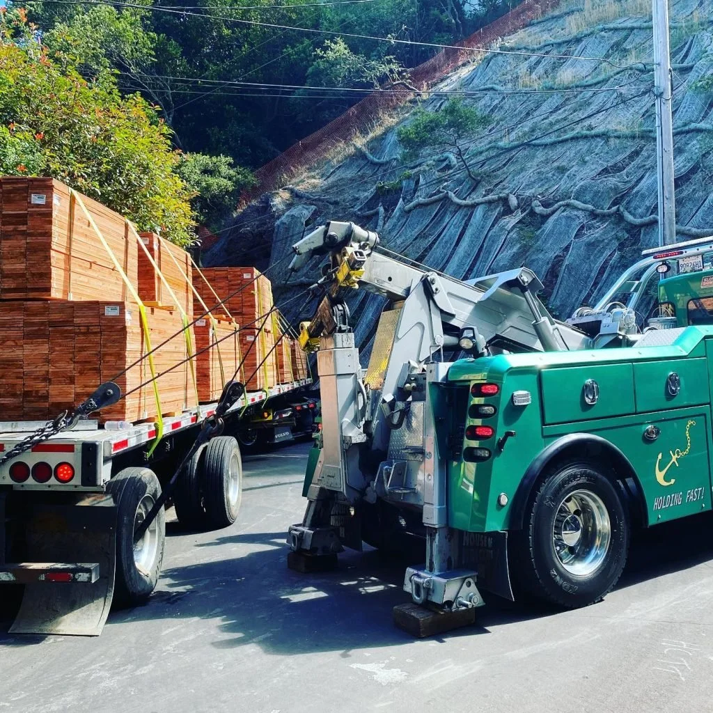 A green tow truck is lifting a flatbed truck that is carrying stacks of lumber, with a rocky hillside and power lines in the background.