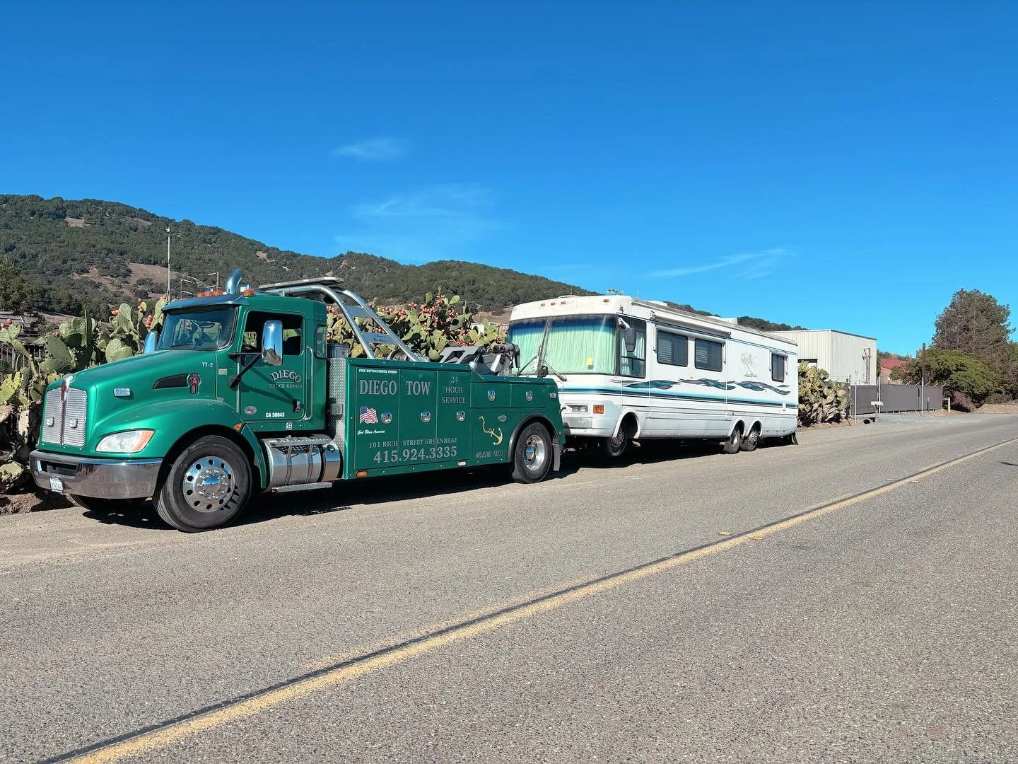 A green tow truck parked on the side of a road next to a white recreational vehicle with cacti in the background and mountains in the distance.