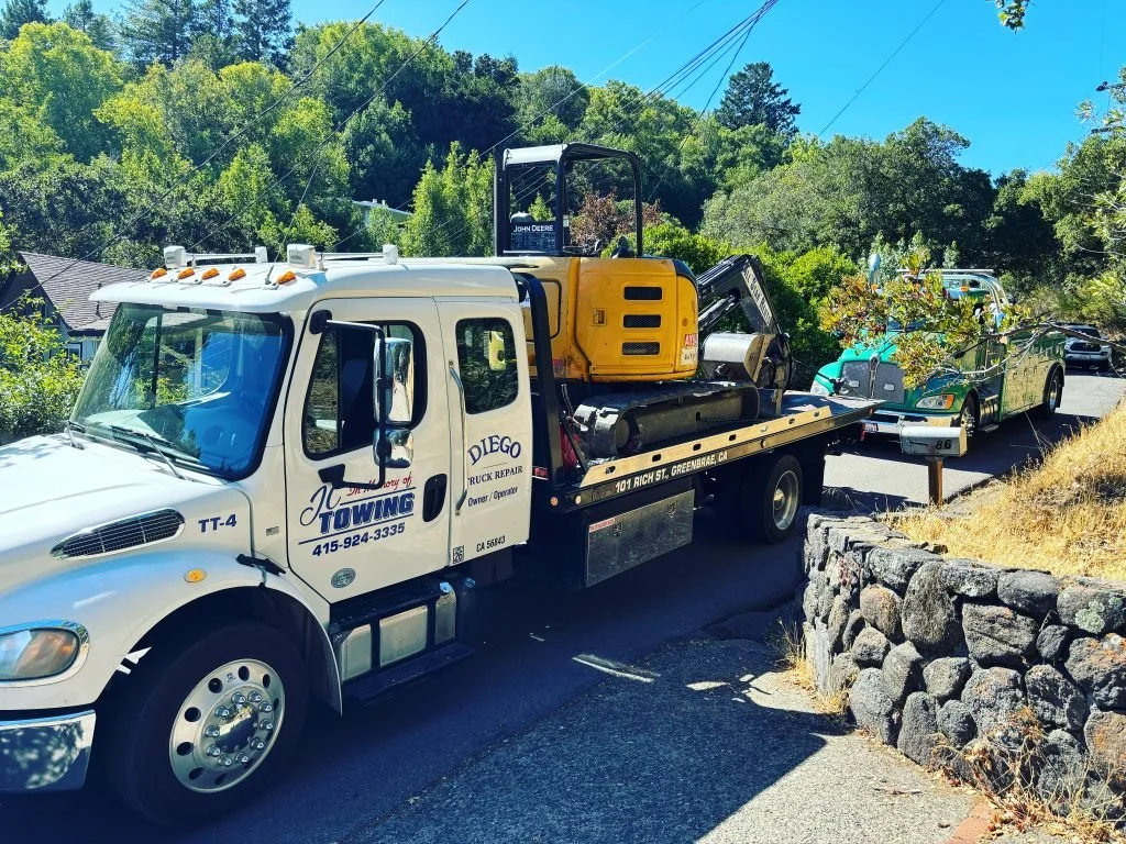 A towing truck with a yellow excavator on its flatbed, parked on a residential street, with two other vehicles behind it and trees in the background.