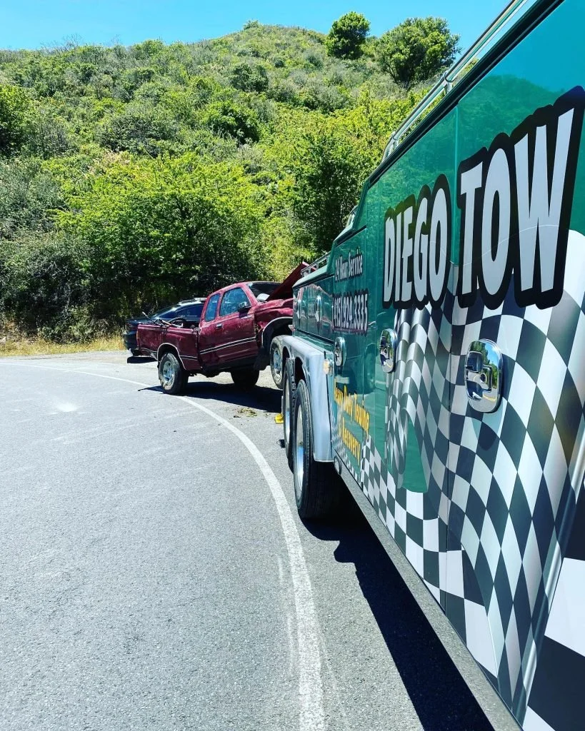 Tow truck from 'Dego Tow' assisting a red pickup truck on the side of a curved road with green trees and hills in the background.