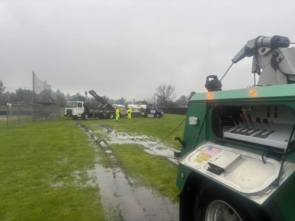 Utility workers on a wet baseball field, with muddy puddles on the grass, repairing or inspecting equipment, amidst overcast weather.