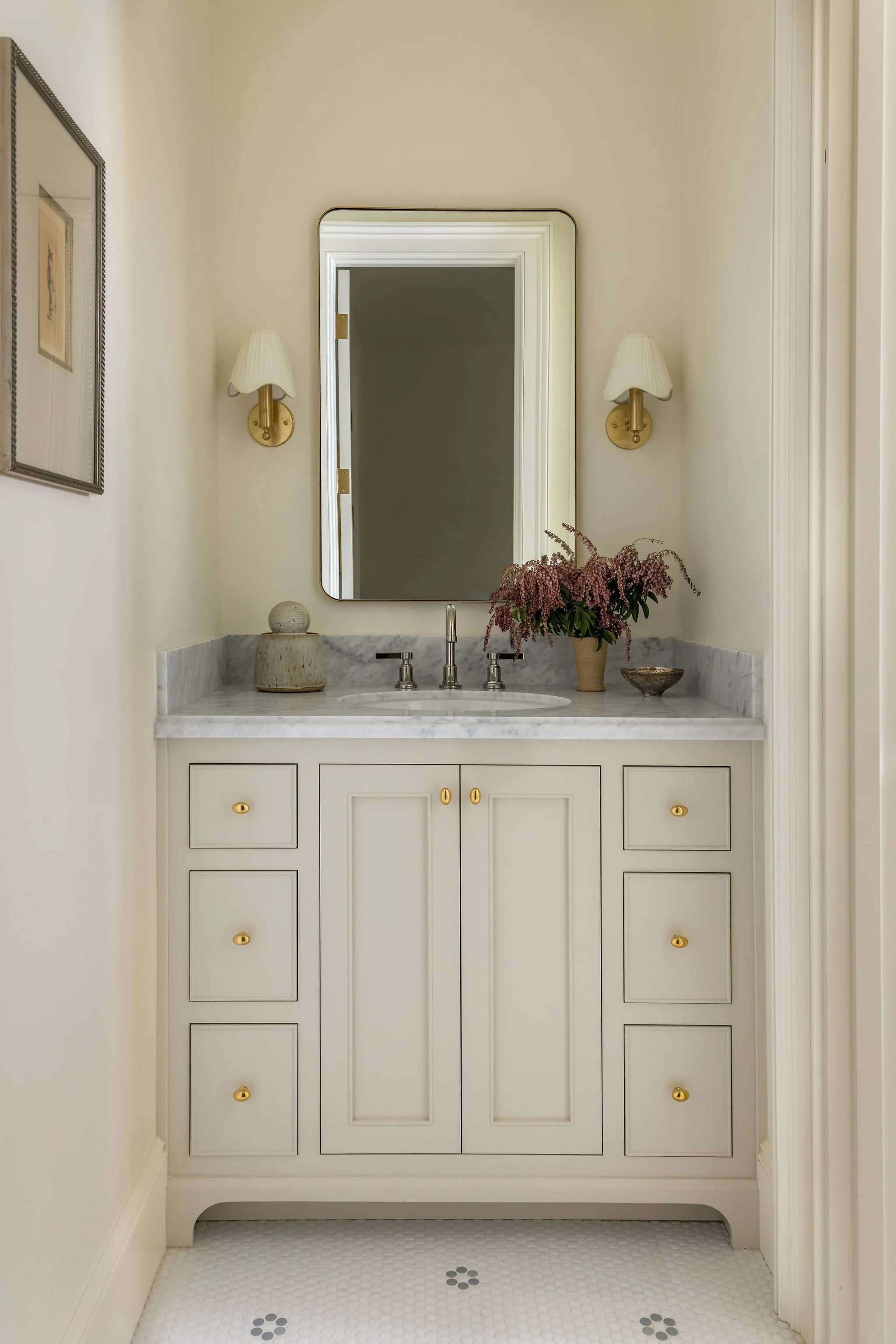 A bathroom vanity with a white marble countertop, a large mirror, two wall sconces with white shades, a potted pink flowering plant, a decorative stone sculpture, and a small stone bowl. The lower cabinet has gold knobs, and the floor is covered with