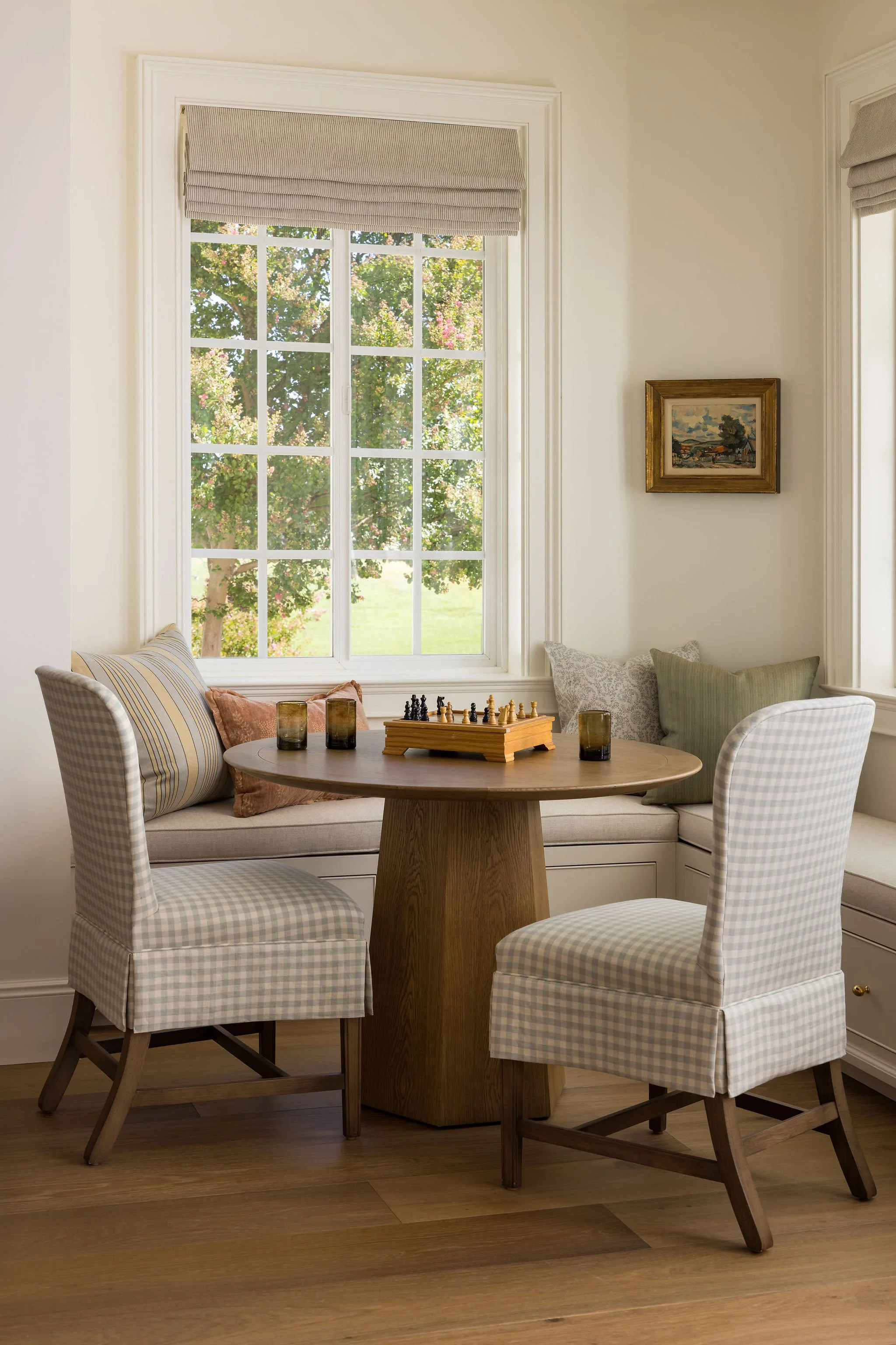 A cozy dining area with a round wooden table surrounded by four checkered upholstered chairs, a window with a view of a lush tree, and a small framed painting on the wall.