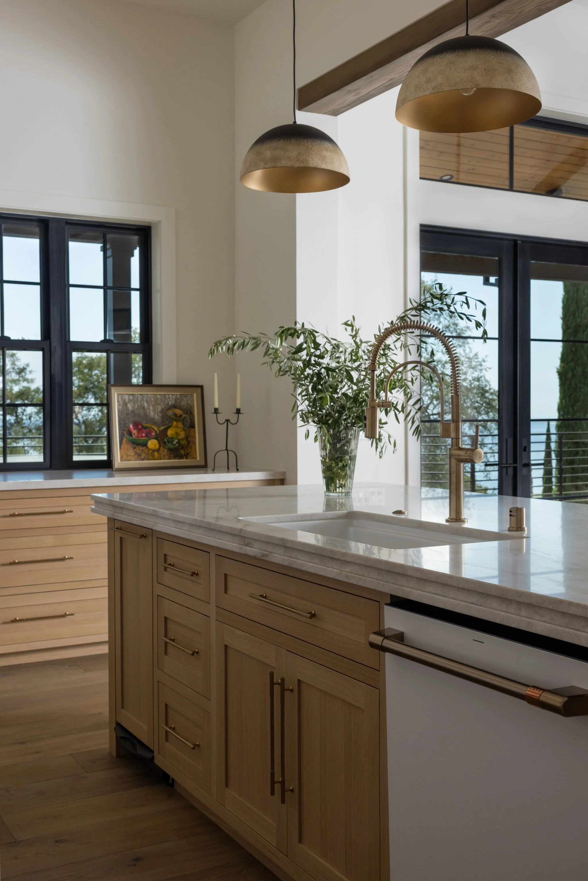 Modern kitchen with a marble countertop island, light wood cabinetry, a gold kitchen faucet, and a vase of greenery. Large windows with black frames and two dome pendant lights hanging from a wooden beam ceiling.