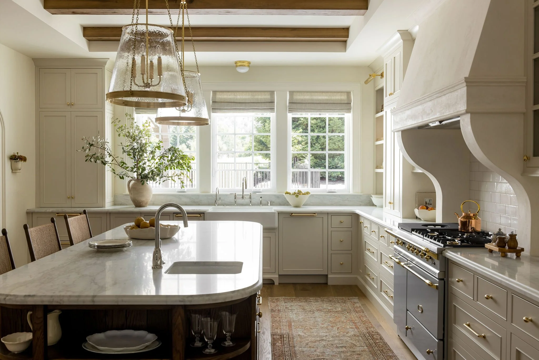 Bright, spacious kitchen with white cabinetry, marble island, large windows, and wooden ceiling beams. A large pendant light hangs over the island, which has a bowl with fruit and plates. There is a plant near the window, and a stove with gold accent