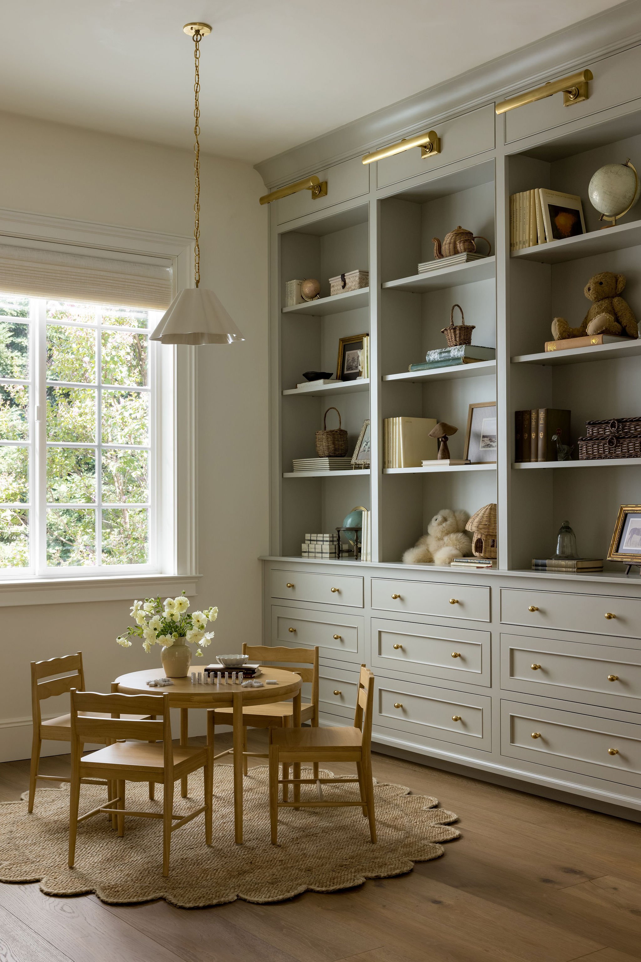 A cozy dining room with a round wooden table surrounded by four wooden chairs, a beige rug underneath, a cream-colored painted built-in cabinet with gold hardware and shelves displaying decorative items, a large window with white trim and blinds, and