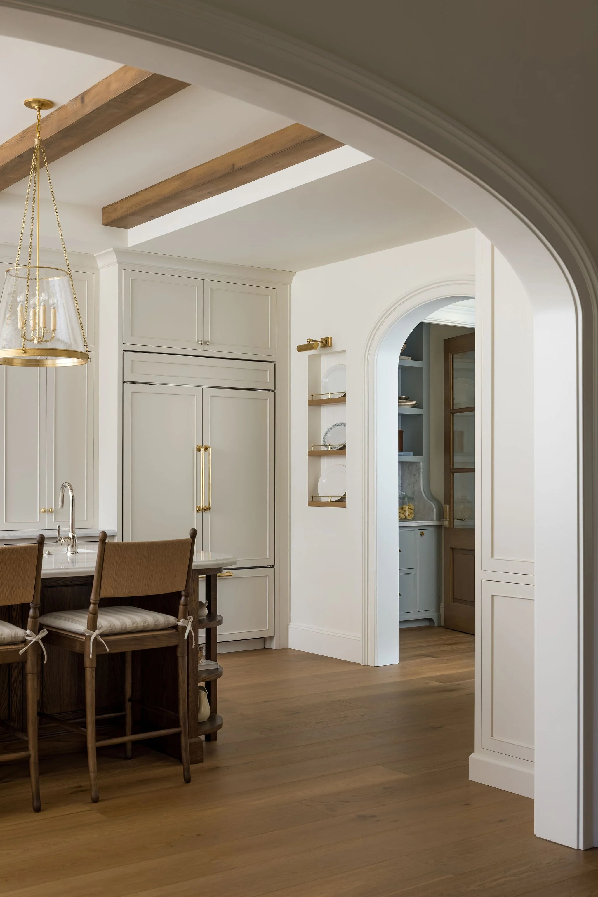 Part of a kitchen or dining area with a wooden table and four chairs, light wood flooring, white cabinetry, a decorative clock, white walls with built-in shelves, and an arched doorway.