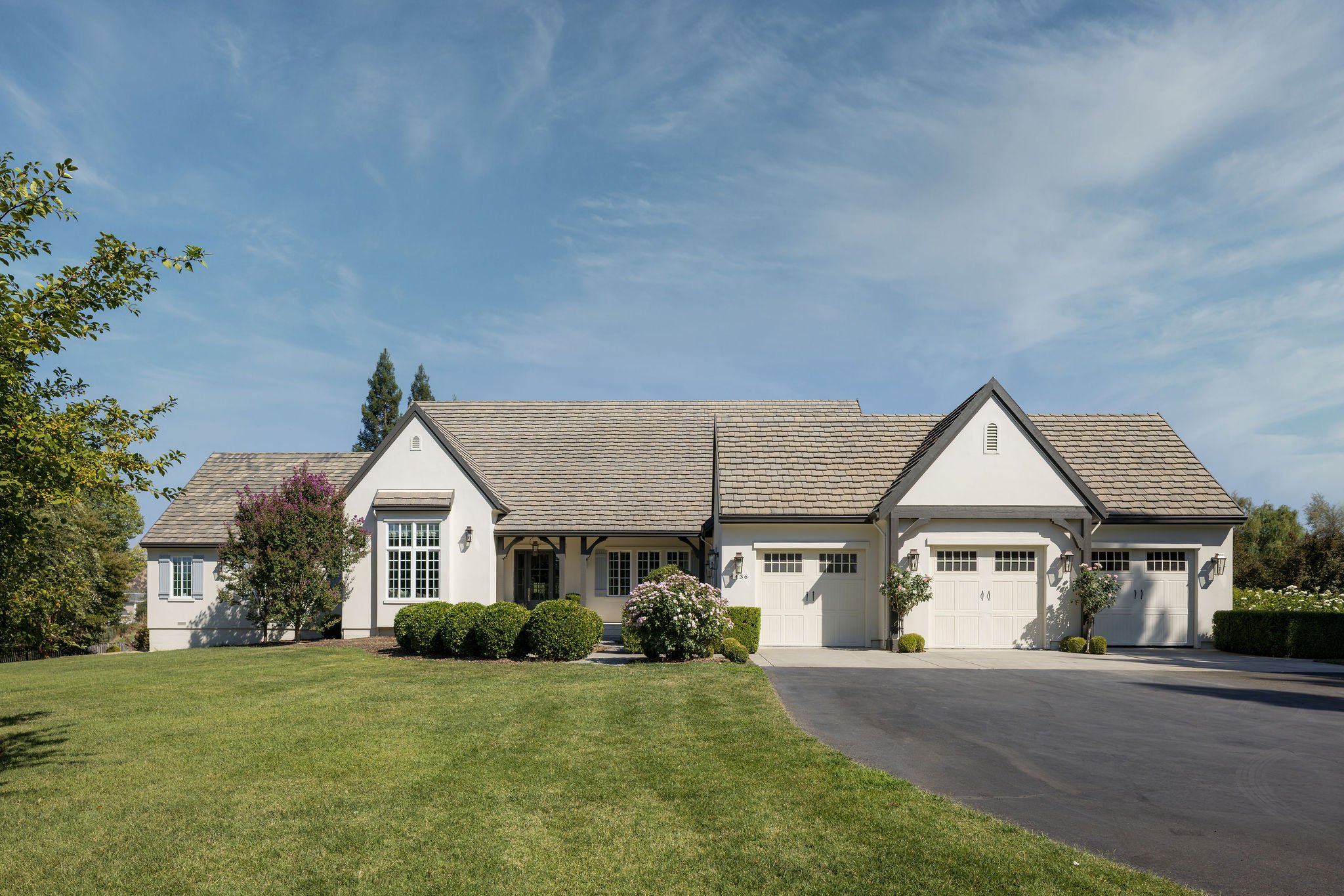 A two-story white house with a gray shingle roof and an attached garage, surrounded by a green lawn, shrubs, and flowering bushes, with a clear blue sky overhead.