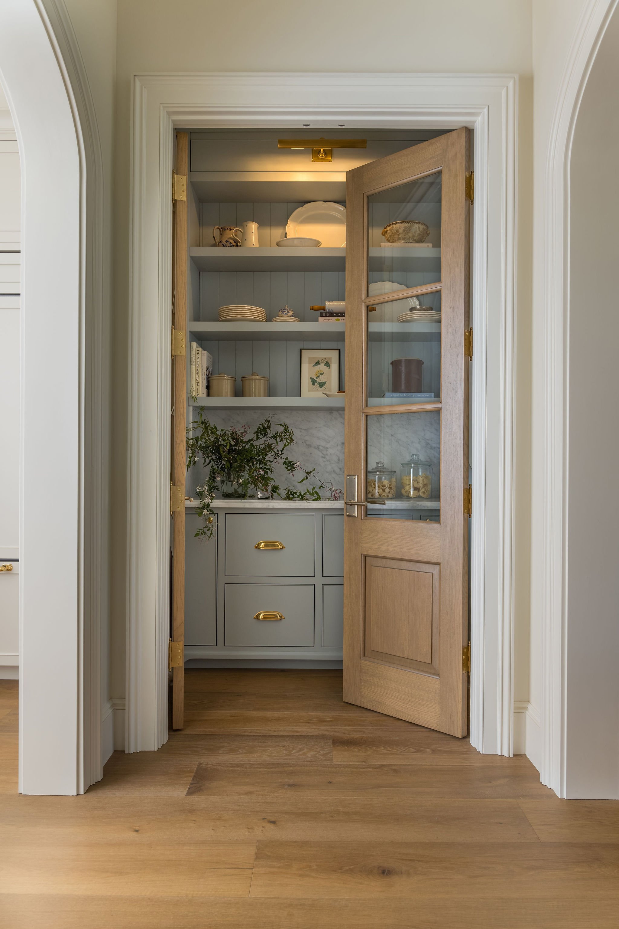 Open wooden cabinet with three shelves containing plates, bowls, and decorative items. The cabinet is set within a doorway with white molding and a marble countertop beneath. The floor is hardwood.