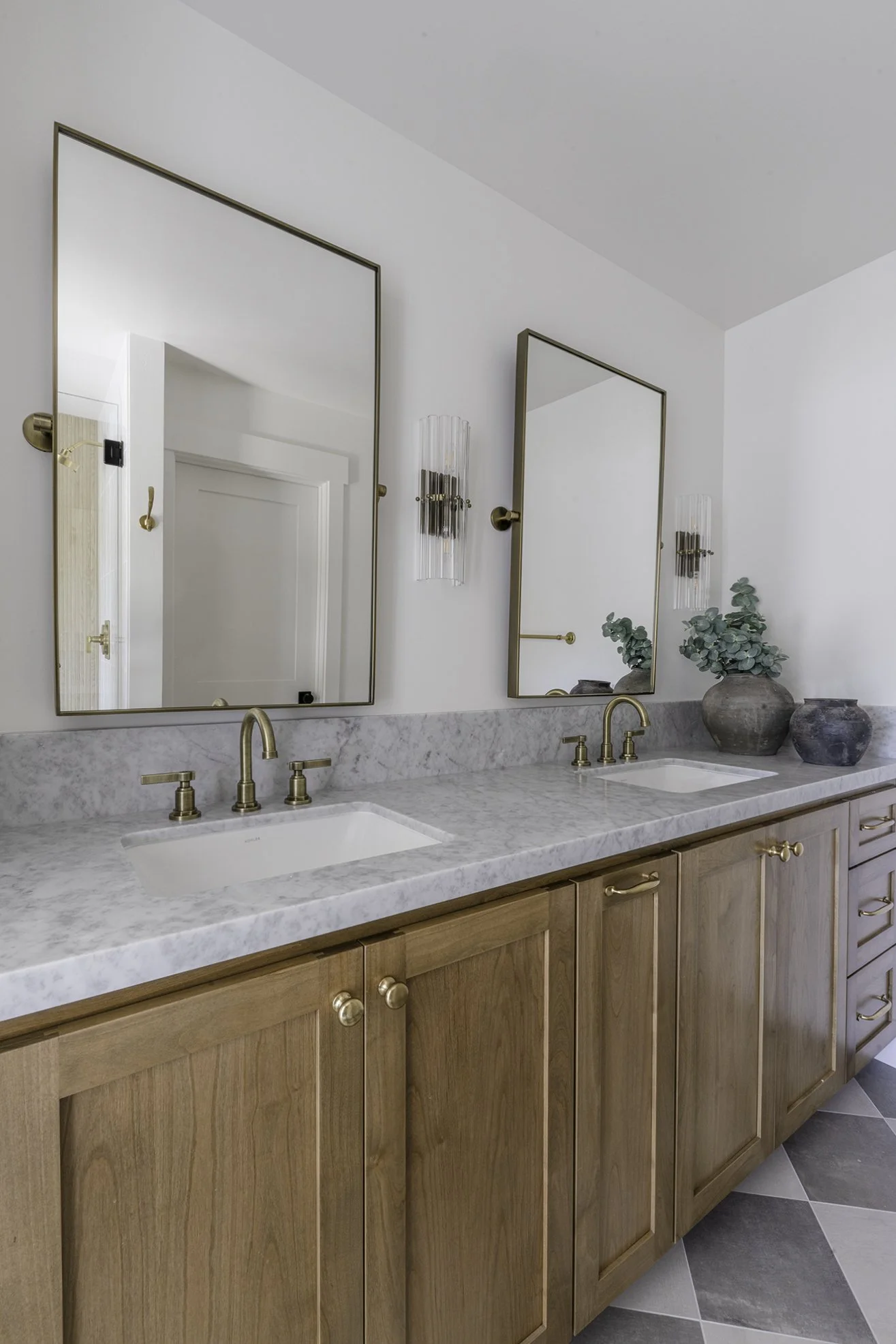 Double bathroom vanity with marble countertop, two rectangular mirrors, wall sconces, wooden cabinetry, and decorative vases with greenery.