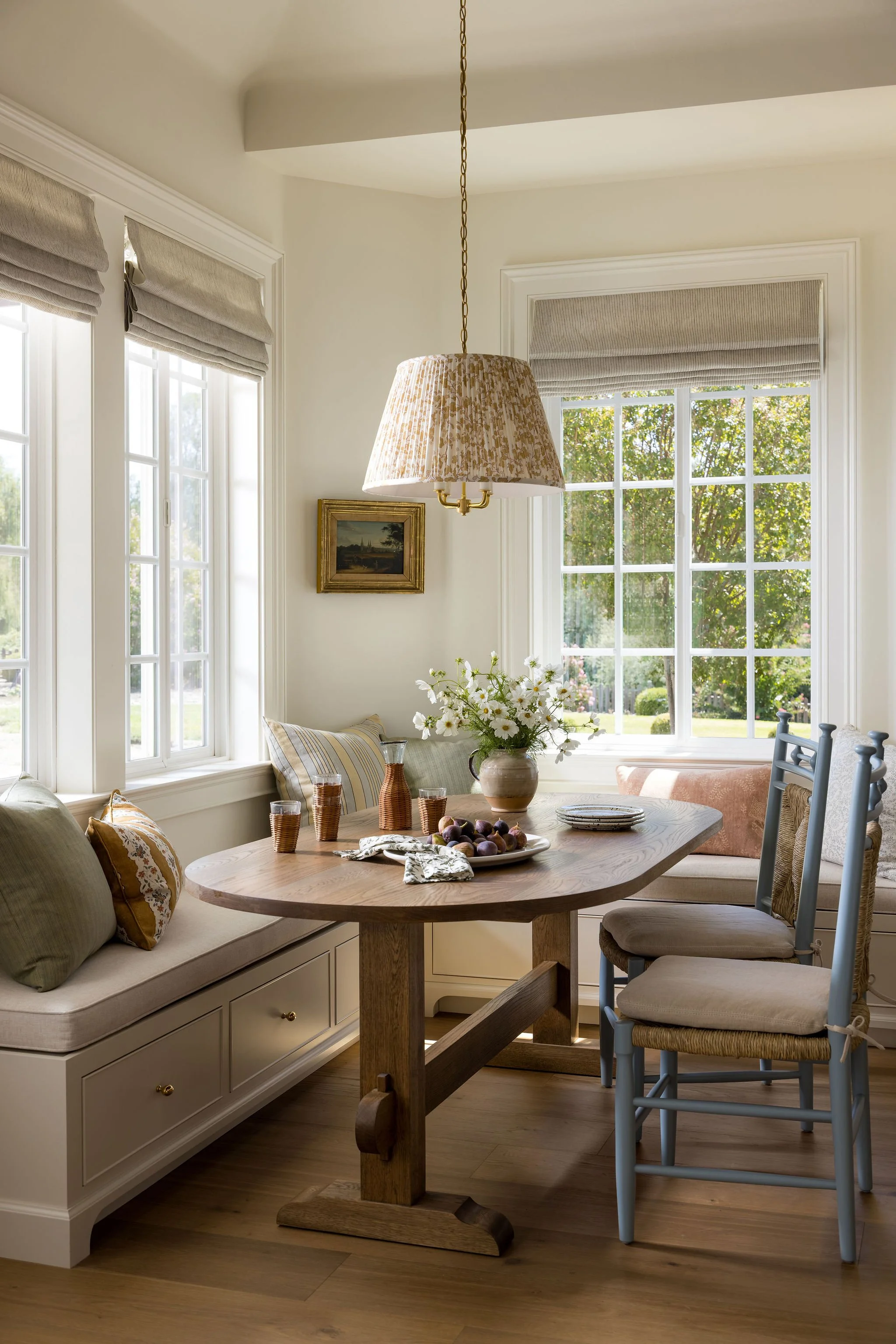 A cozy dining area with large windows, beige window blinds, a tree-lined view outside, and a round wooden table set with a vase of white flowers, a plate of grapes, and glasses. There is a built-in white bench with cushions and pillows, and four whit