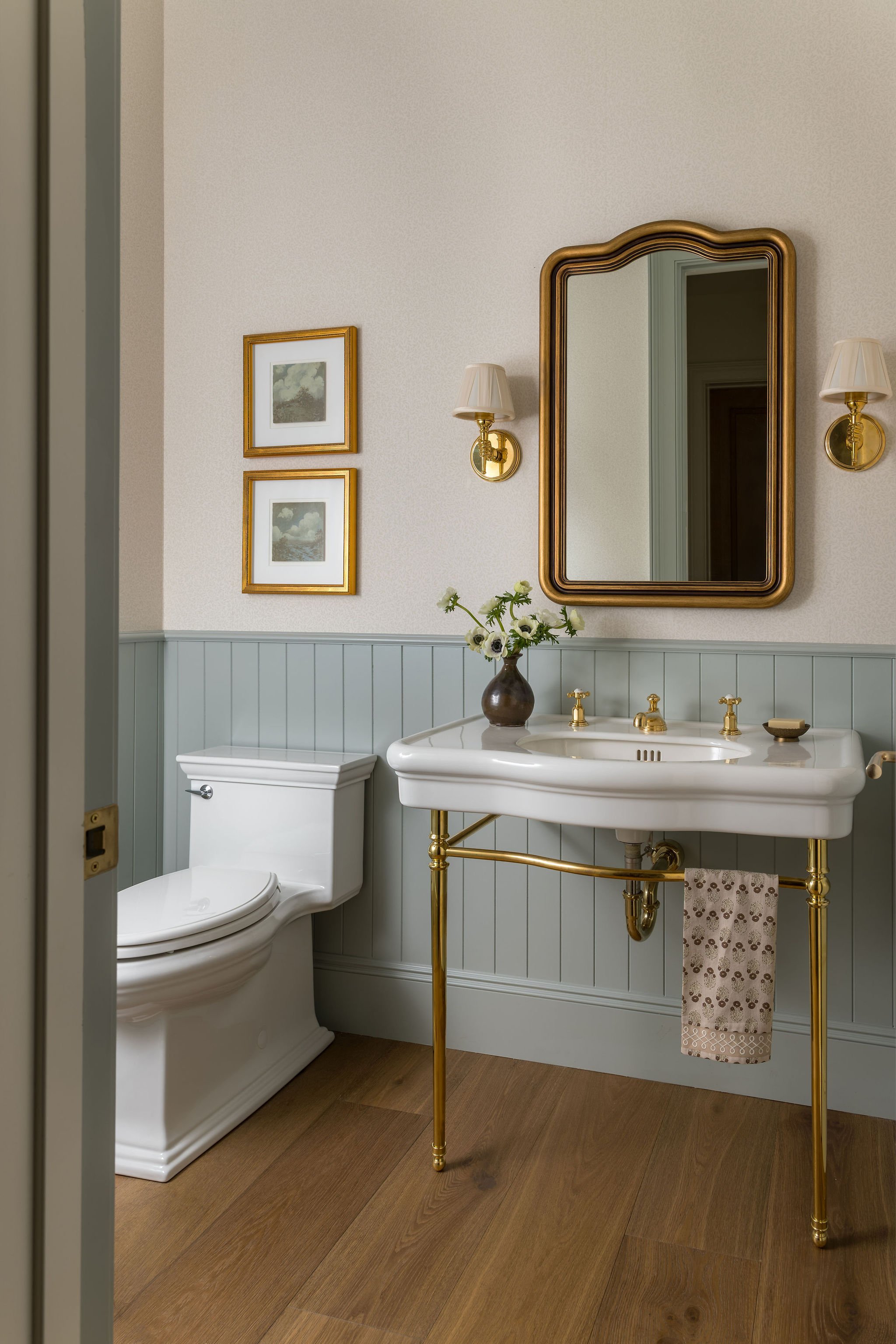 A bathroom with a white toilet next to a gold and white pedestal sink, a large mirror with a gold frame, two wall-mounted light fixtures, framed artwork on the wall, and a small vase with white flowers on the sink.