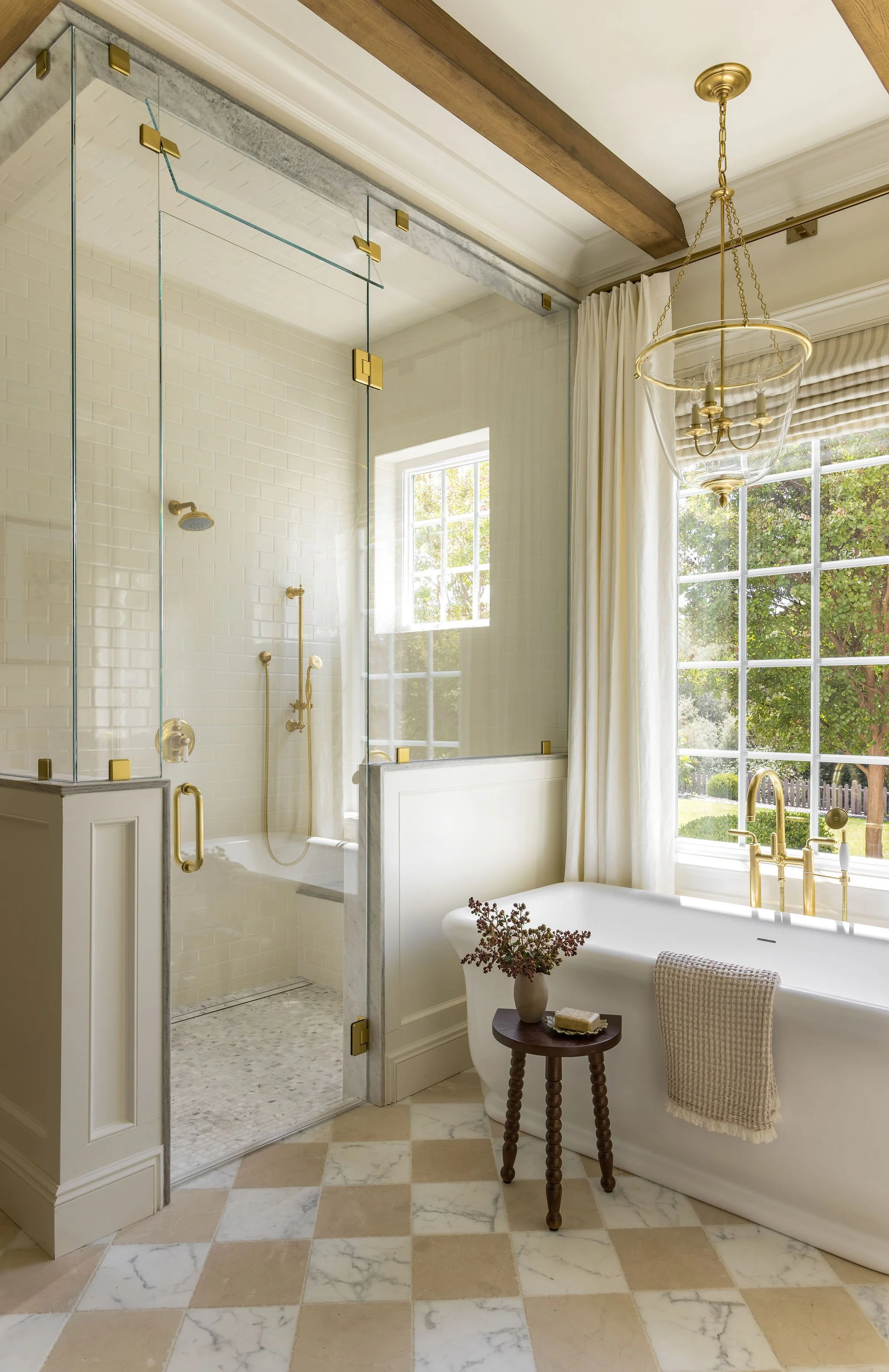A bathroom with a walk-in shower, clawfoot bathtub, large window with curtains, chandelier, and checkered floor tiles.