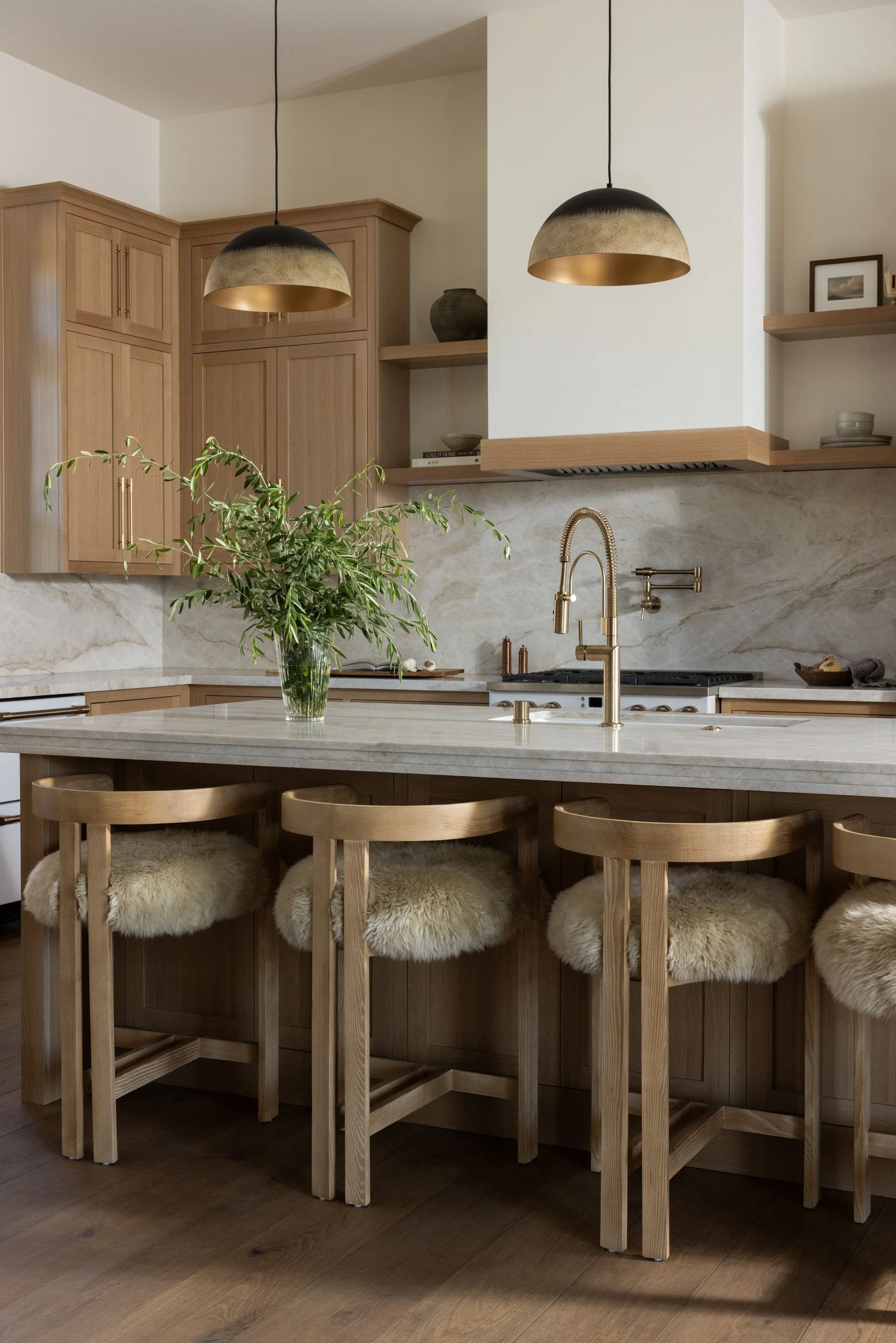 A modern kitchen with wooden cabinetry, a marble island, and a row of four wooden bar stools with fur-covered cushions.