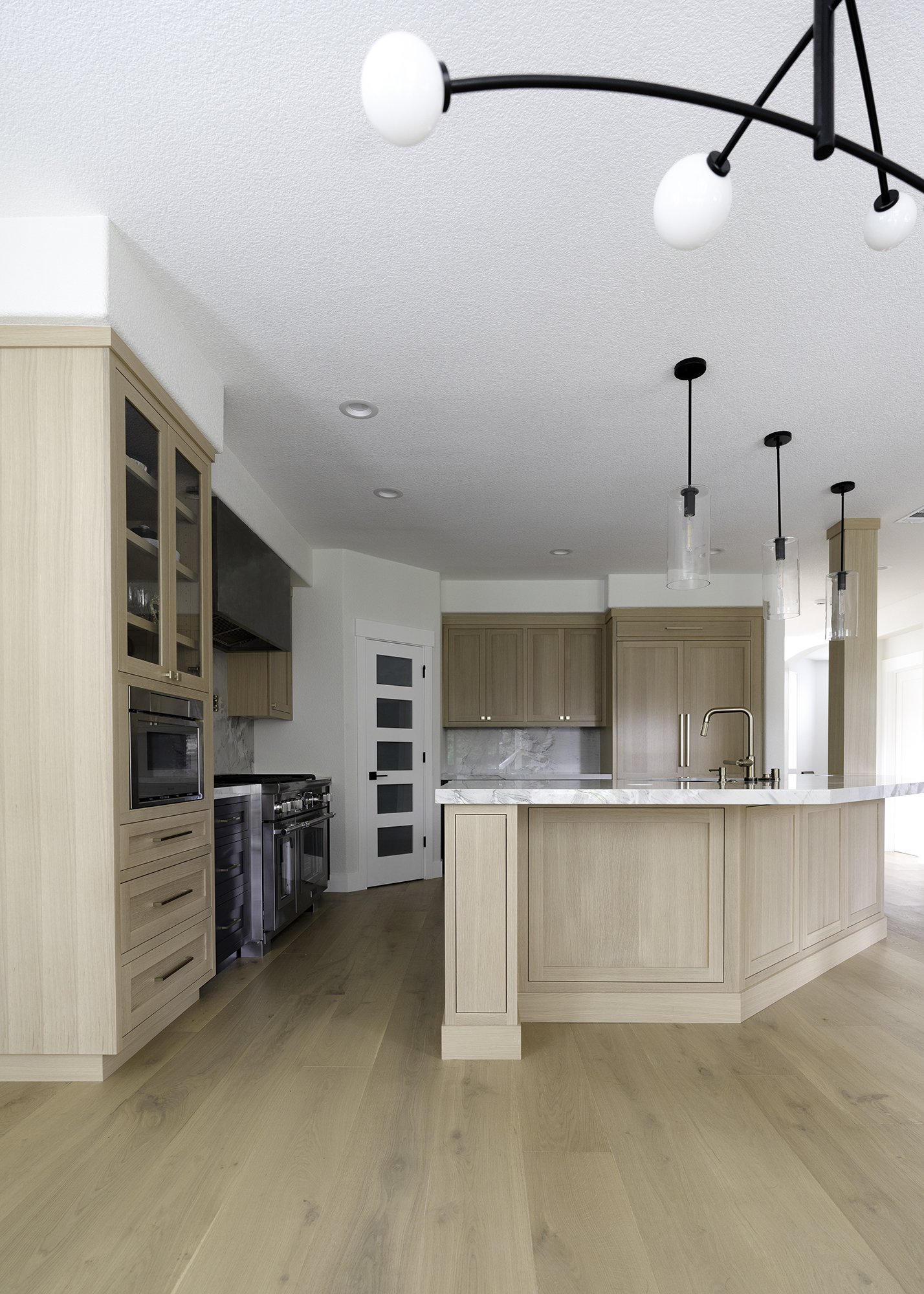 Modern kitchen with light wood cabinets, marble countertops, and a kitchen island with a sink. Features pendant lighting and a ceiling fixture with multiple bulbs, hardwood flooring, and a white door with glass panels.