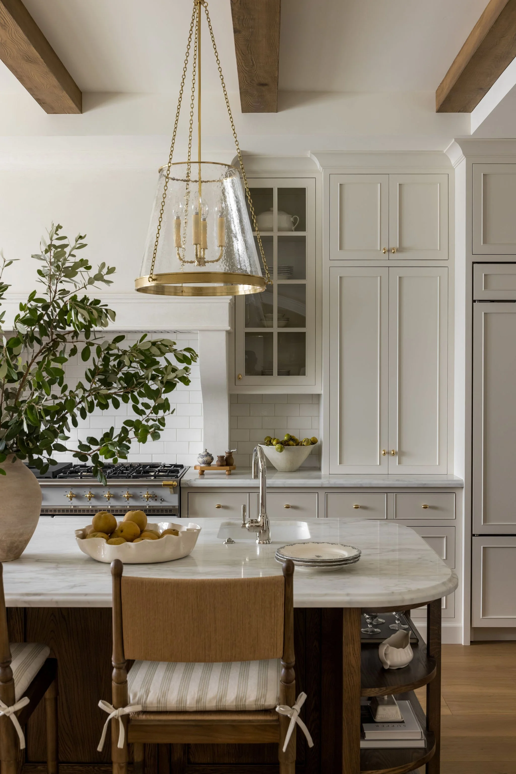 A kitchen with white cabinets, a marble countertop, a country-style wooden table with chairs, a large beige bowl with pears, a gold chandelier, and a green plant.