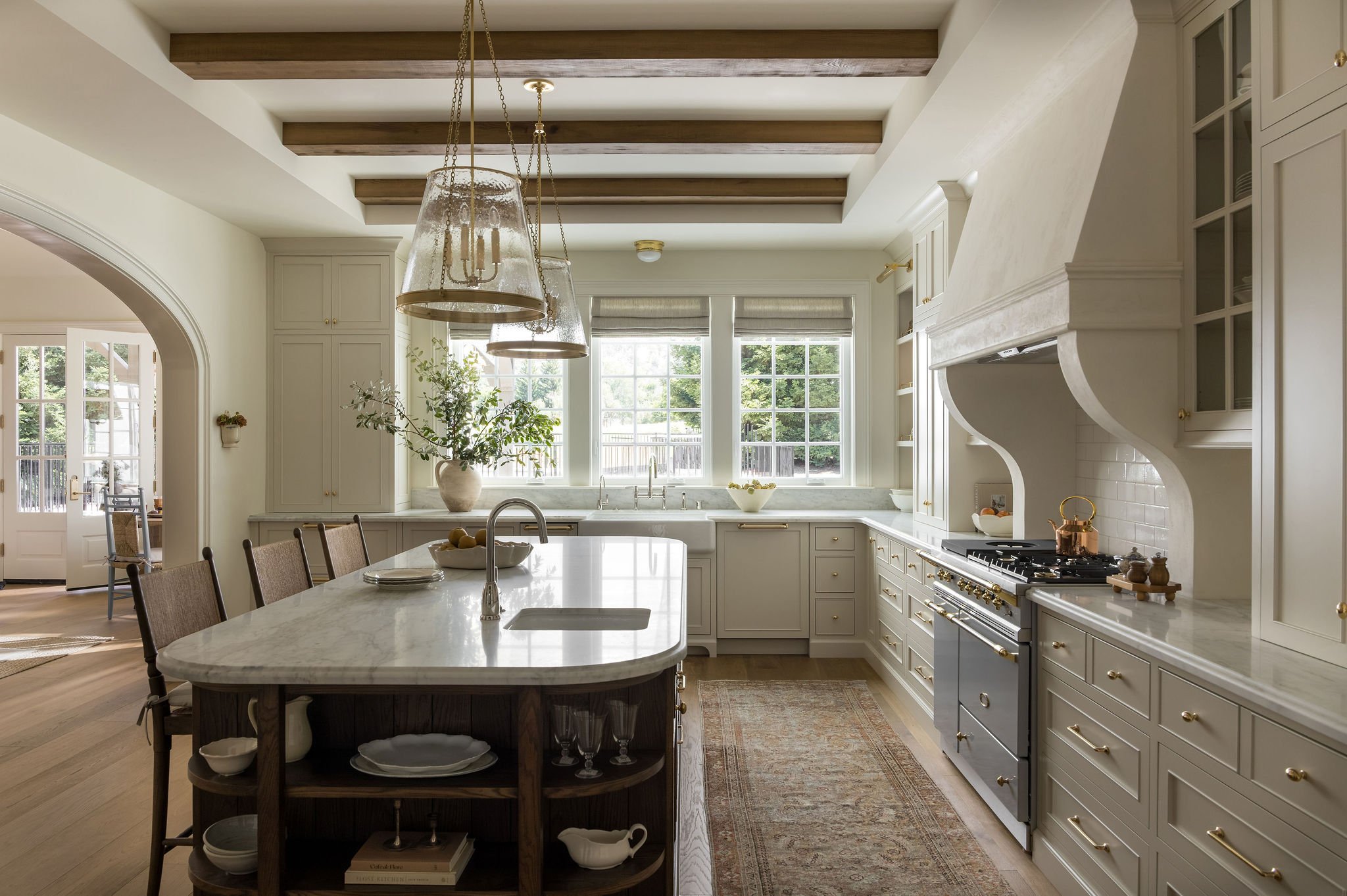 Bright kitchen with white cabinets, large window, and a kitchen island with a white marble countertop, decorated with a bowl of fruit, on a hardwood floor, and a wooden chandelier hanging from the ceiling.