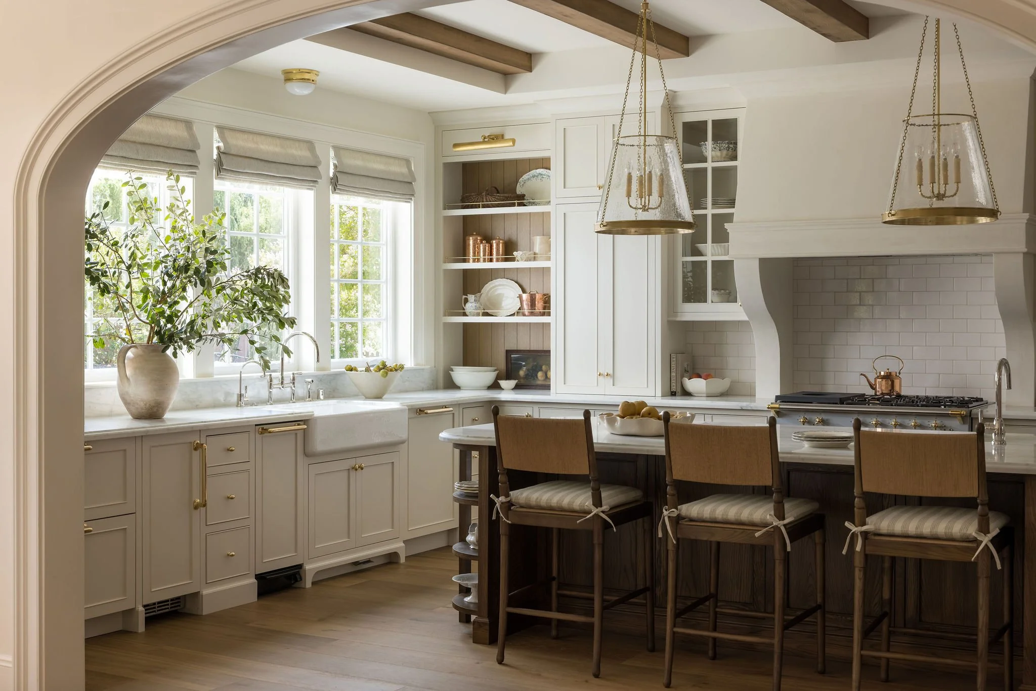 A cozy kitchen with white cabinets, wooden beams on the ceiling, and a large window with Roman shades. There's a marble countertop with a potted plant, a bowl of lemons, and a farmhouse sink. A built-in open shelf displays dishes and copper cookware.