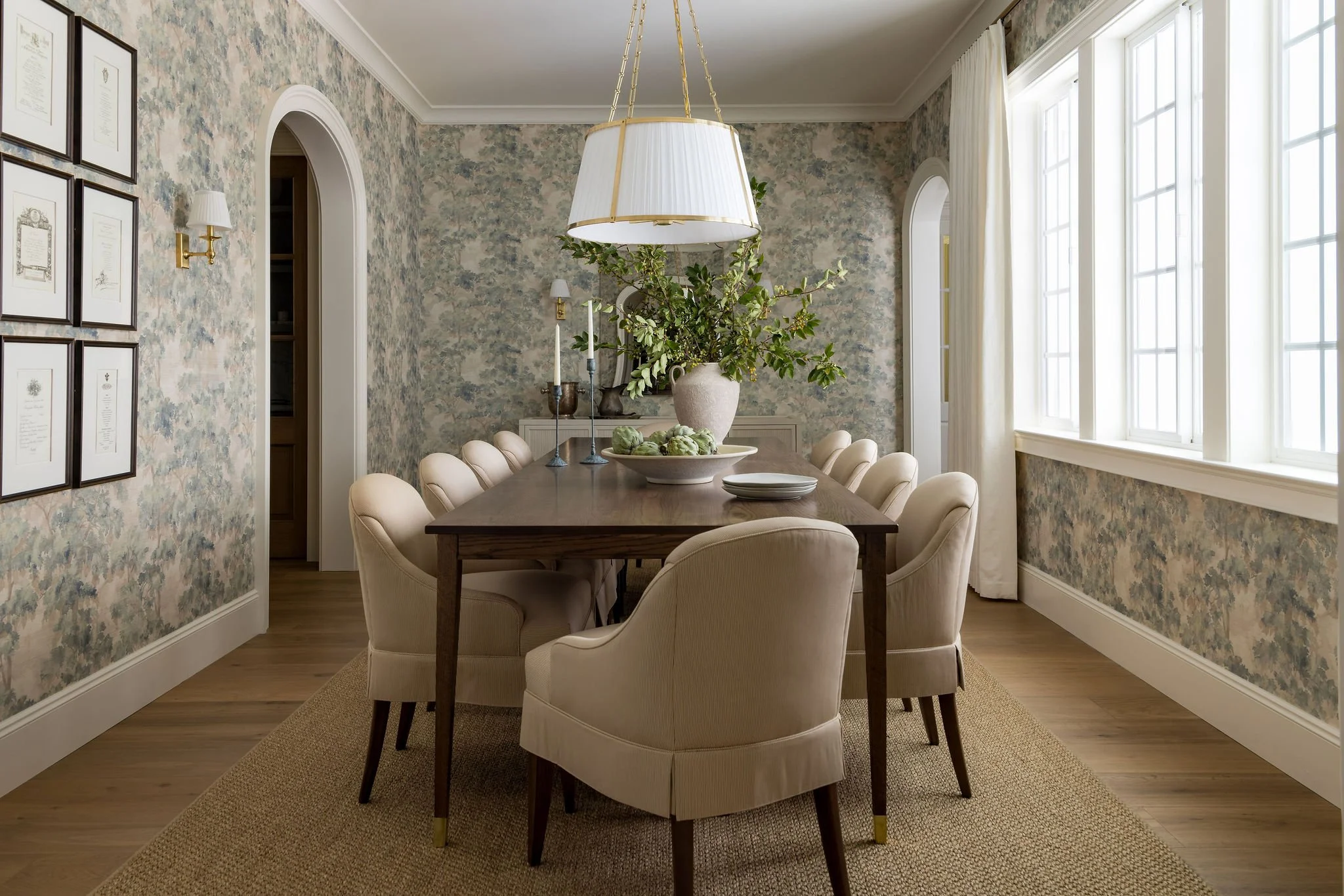 A cozy dining room with a wooden table surrounded by cream upholstered chairs. The table features a large vase with green foliage, a bowl of artichokes, and candlesticks. The room has floral wallpaper, framed documents on one wall, and large windows 