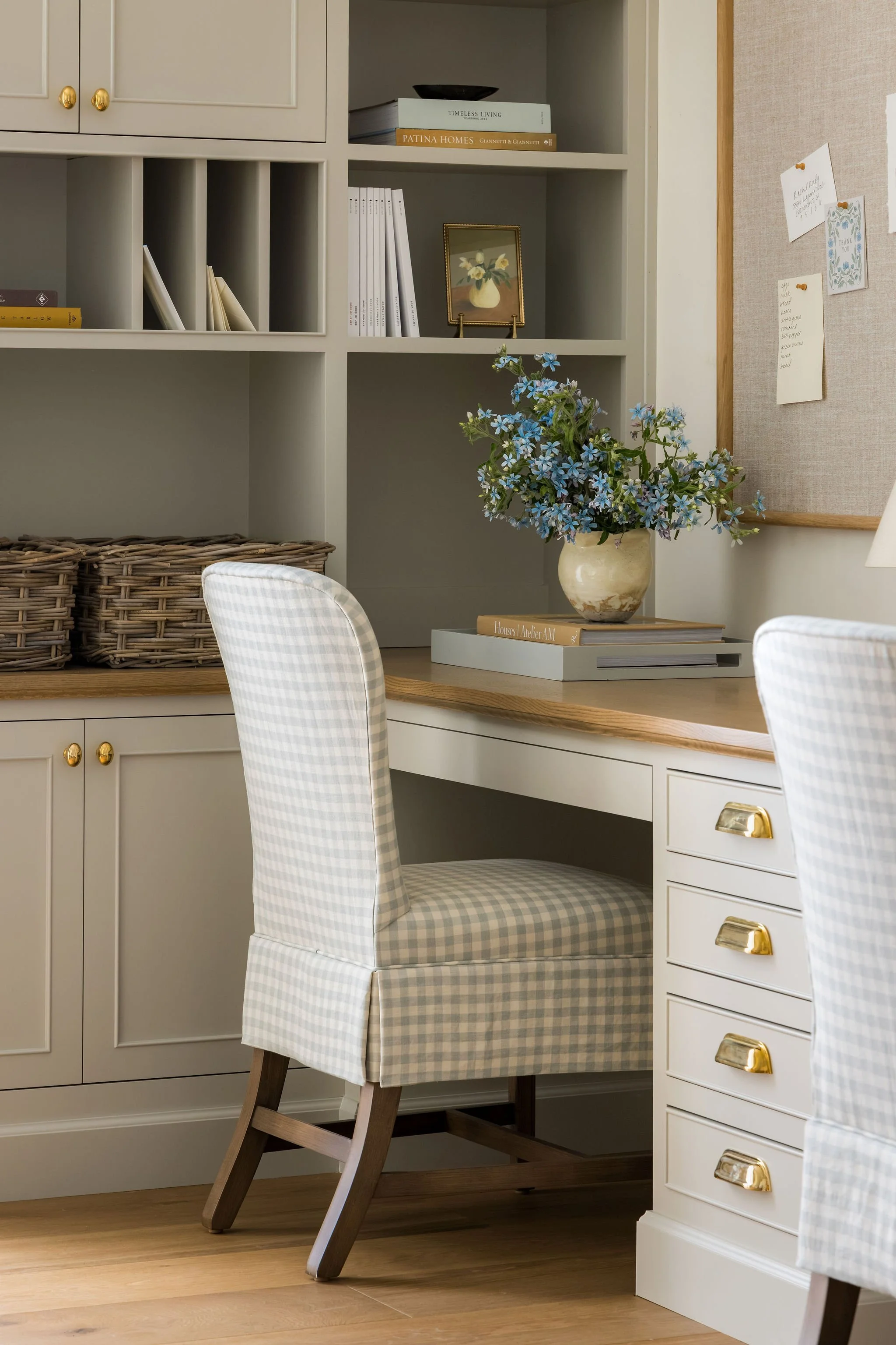 A home office or study area featuring a light-colored desk with gold hardware drawers, a plaid upholstered chair, and a flower arrangement with blue flowers in a cream-colored vase. Behind the desk is a white built-in bookshelf with books, a small fr