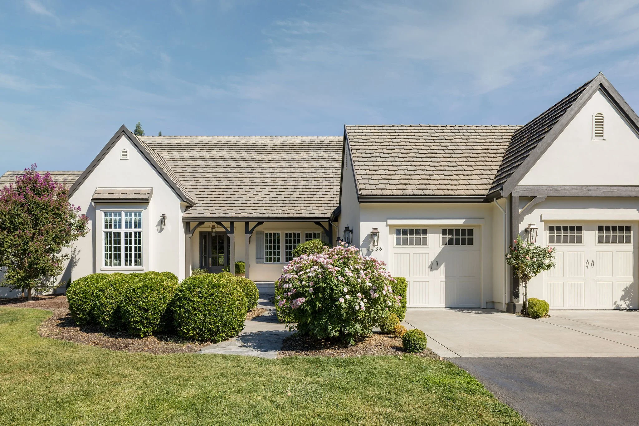 Front view of a white two-story house with a gray shingle roof, white garage door, and a front yard with bushes and flowering plants.