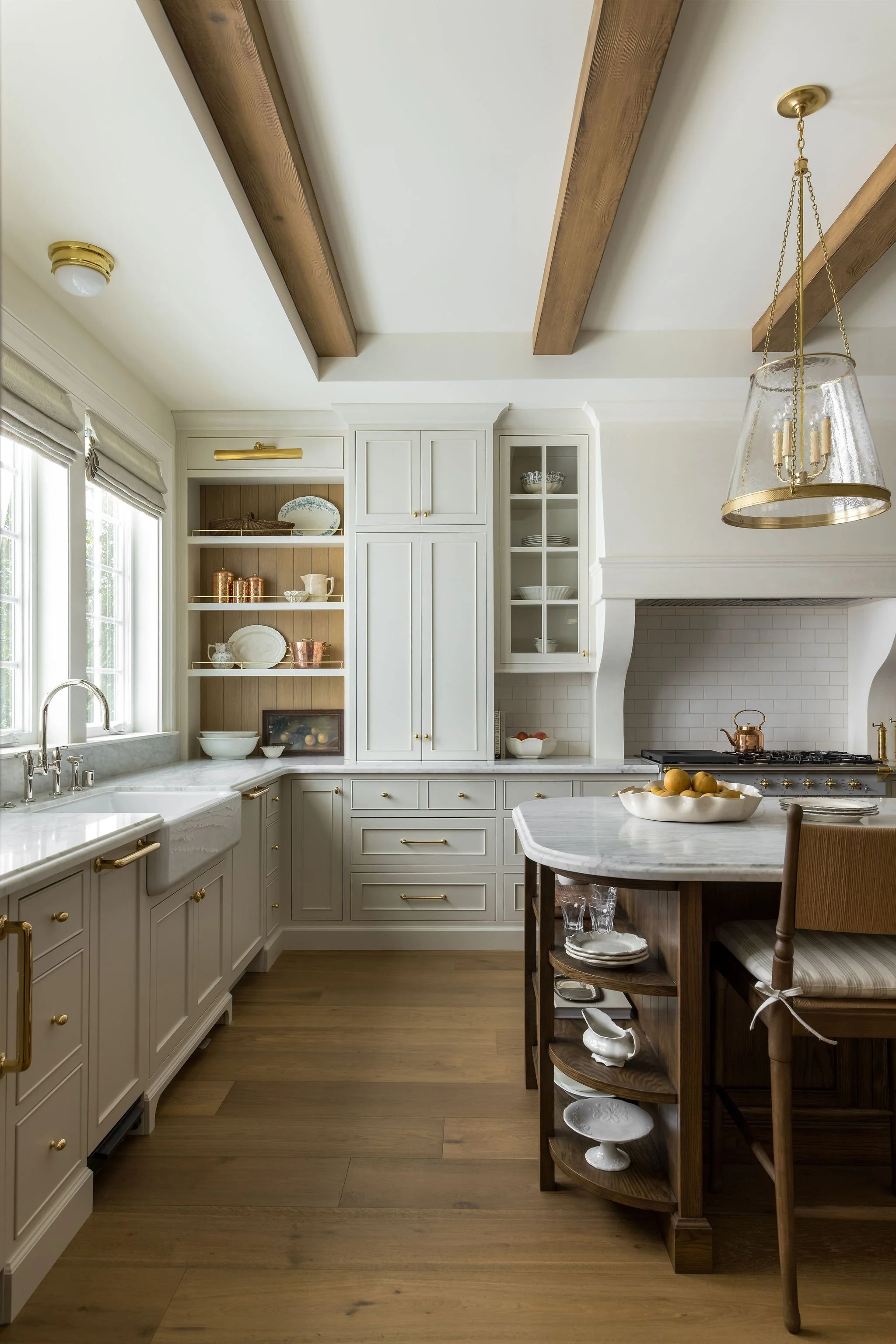 A kitchen with white cabinets, a marble countertop, a wood kitchen island with a marble top, and wooden beams on the ceiling. There is a hanging light fixture with brass accents and a cluster of lemons in a white bowl on the kitchen island. The kitch