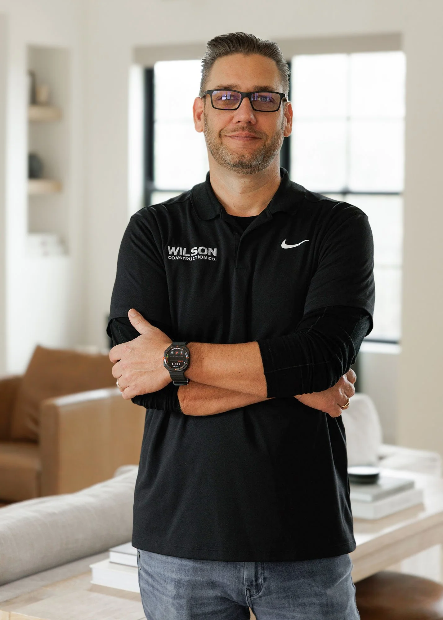 A man with glasses wearing a black polo shirt with "WILSON CONSTRUCTION CO." and Nike logo, standing indoors with arms crossed, in a room with large windows and modern decor.