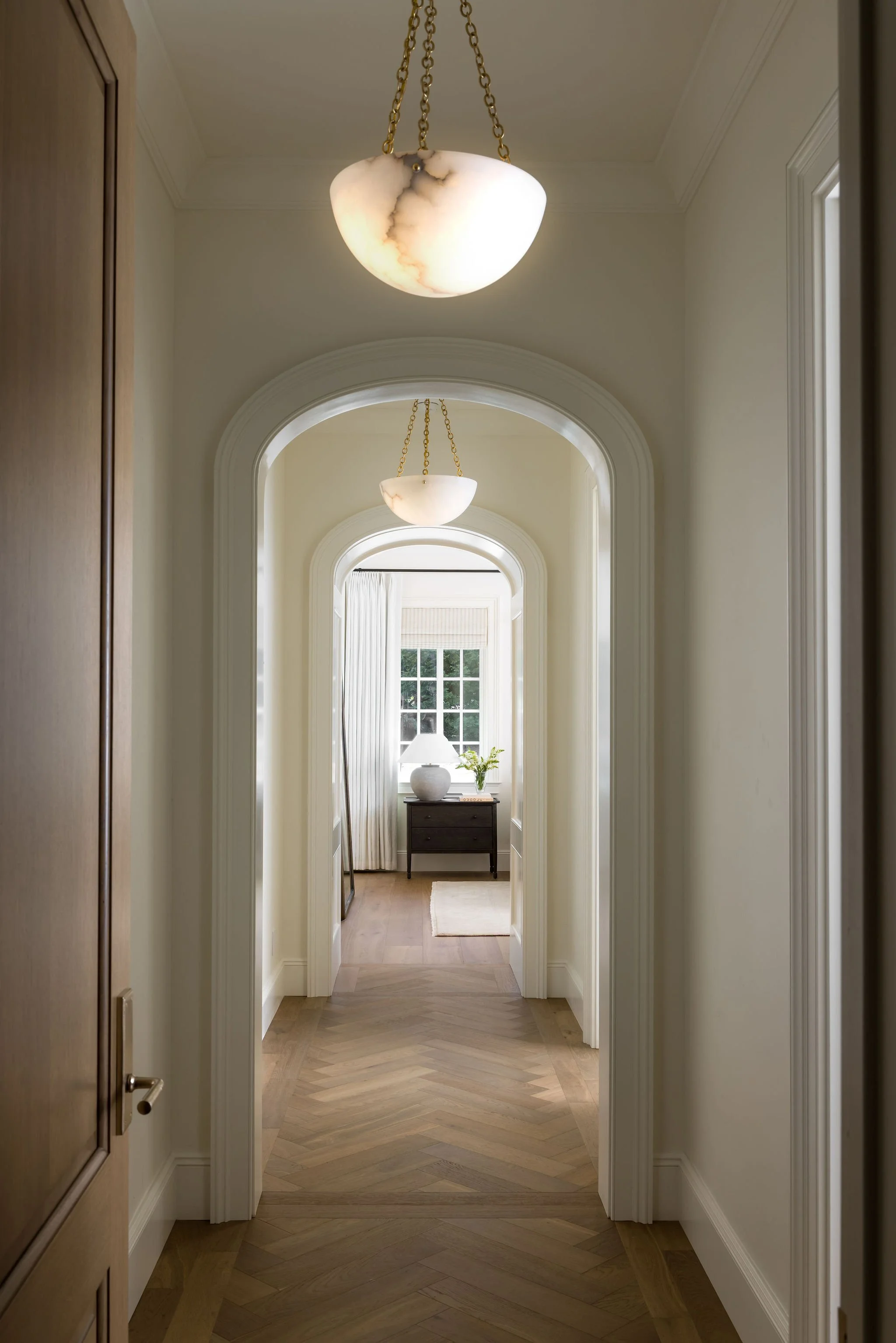 A hallway with arched doorways and a large window at the end, decorated with a table, lamp, and potted plant, with hanging lantern-style light fixtures on the ceiling.