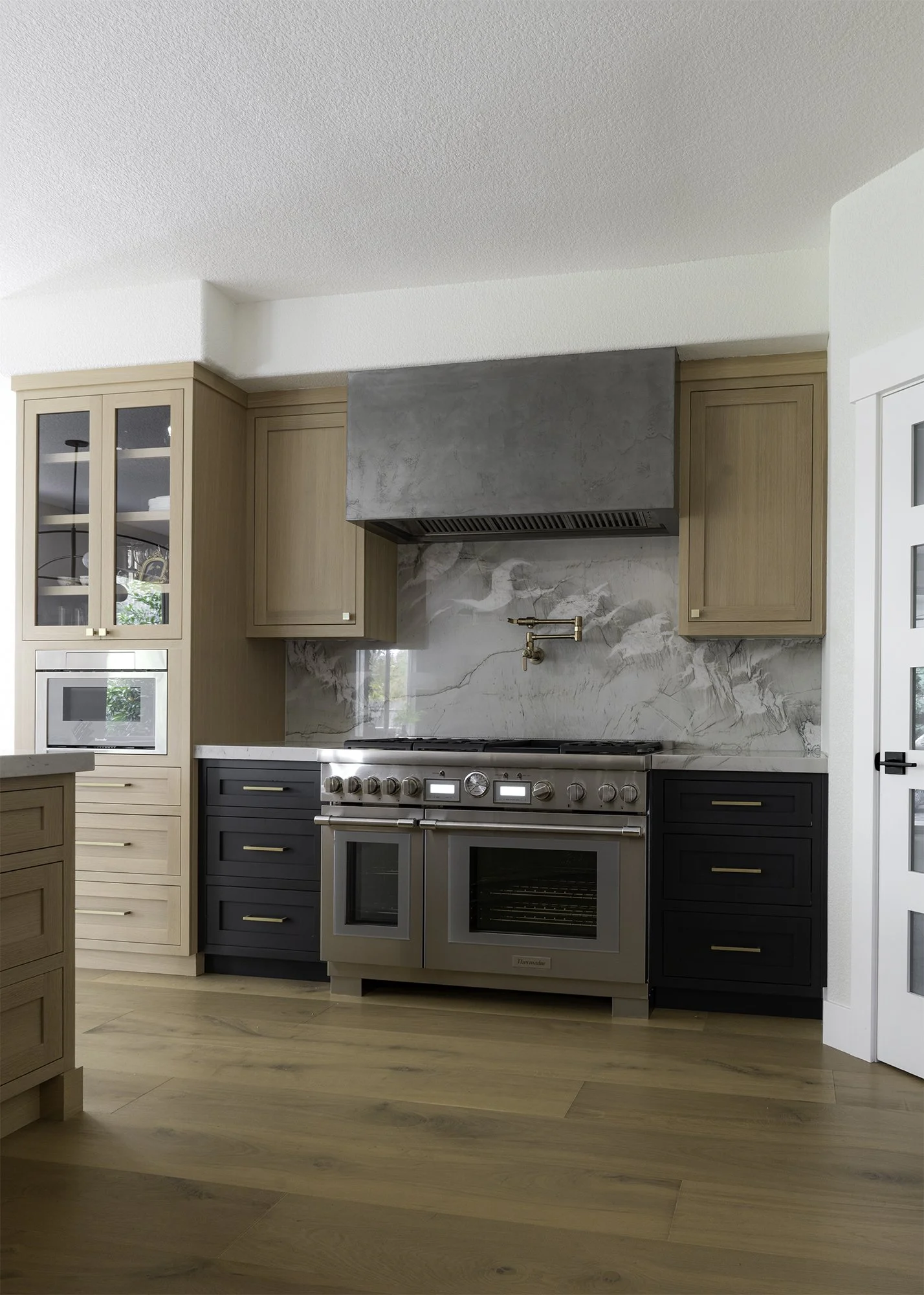 Modern kitchen with wooden cabinetry, stainless steel range with double ovens, marble backsplash, and a large range hood. Light wood flooring and a glass-front cabinet visible.