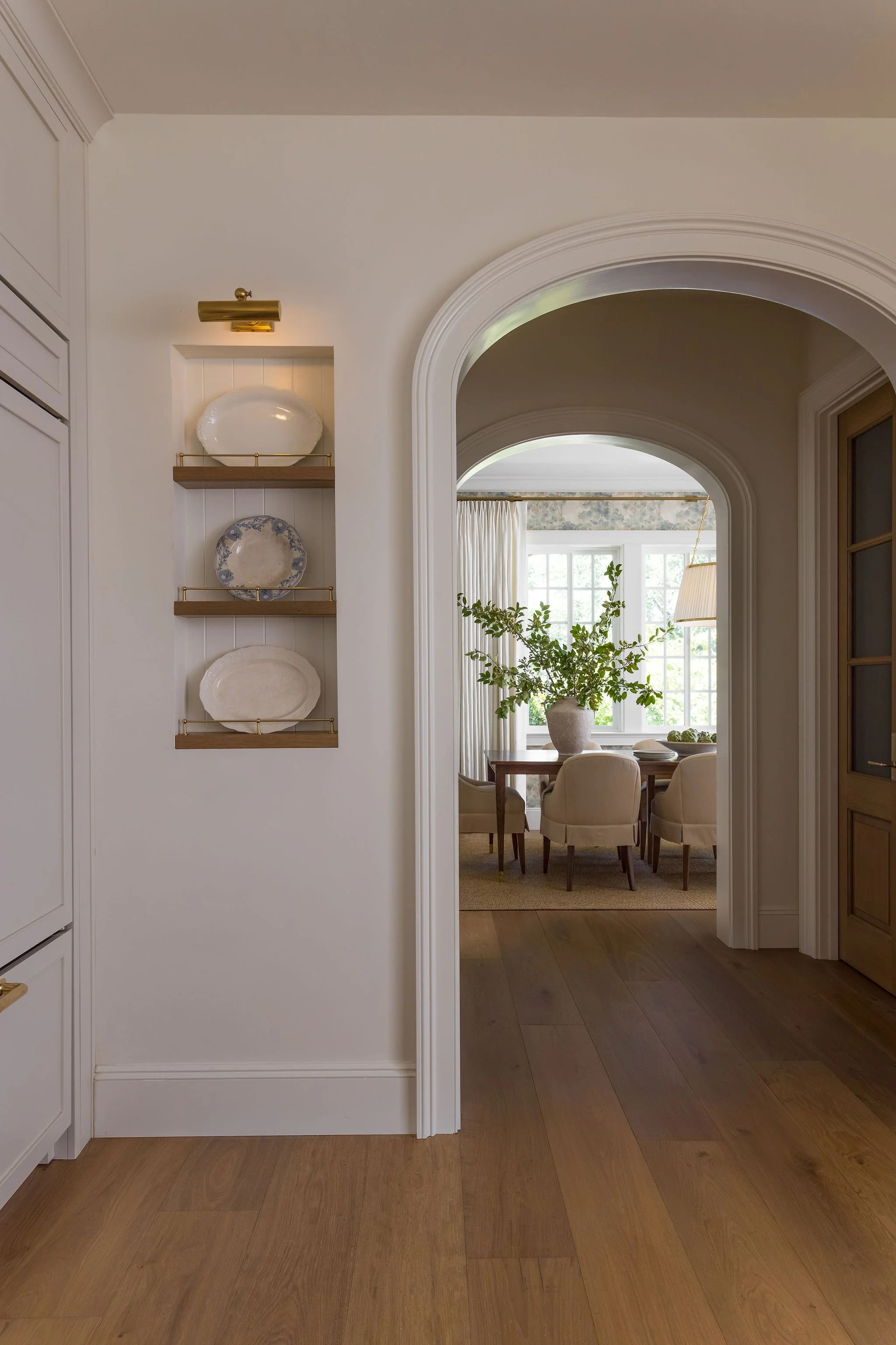 View through an arched doorway into a dining area with a round wooden table, beige upholstered chairs, a large window with curtains, a potted plant, and a hanging lamp.