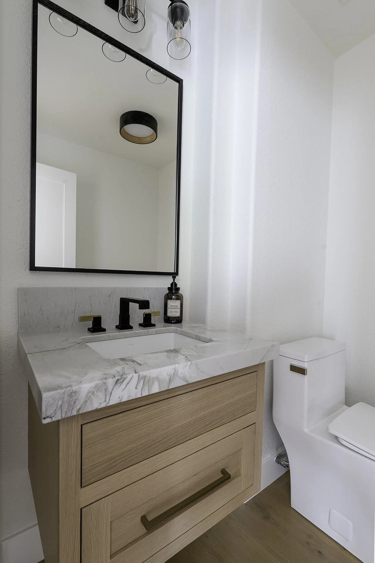 Bathroom vanity with a marble countertop, black faucet, brown wooden cabinet, a large wall mirror, a soap dispenser, and a white toilet, with a modern ceiling light fixture reflected in the mirror.