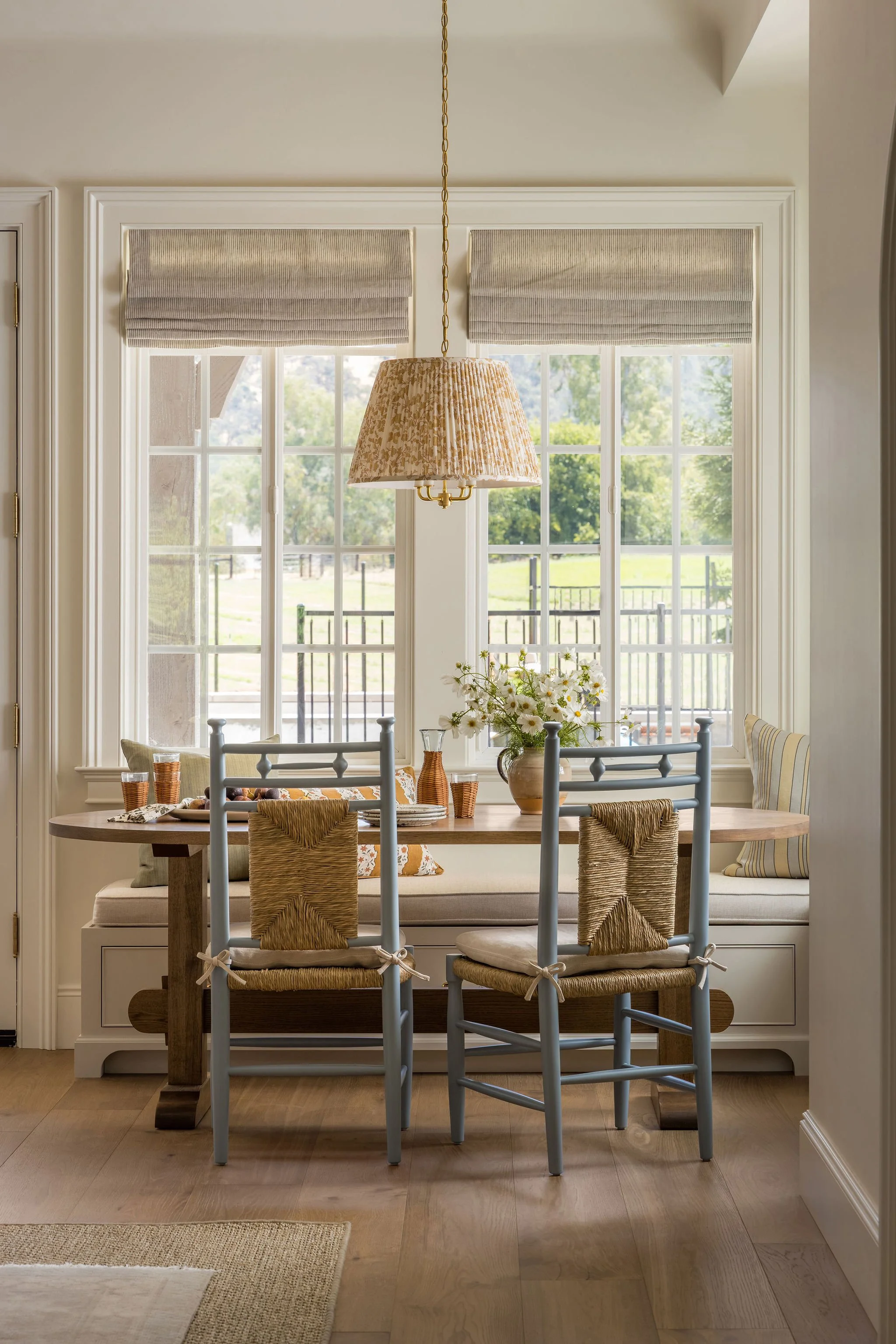 A sunlit dining area with a window seat and a round wooden table surrounded by six chairs. The window has two shades, and the wall has a hanging pendant light with a patterned lampshade. There are decorative pillows and a flower arrangement on the wi