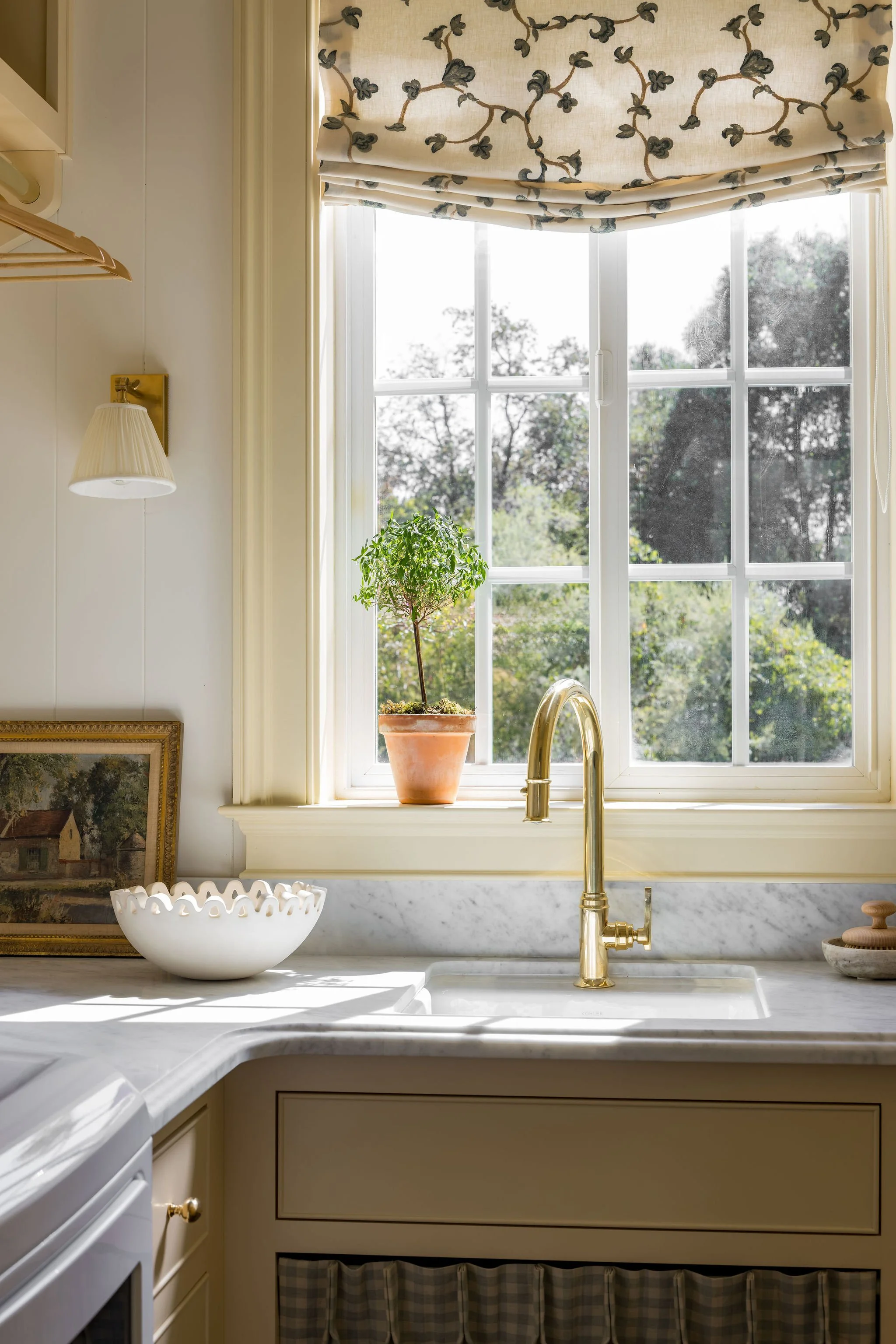 A bright kitchen window with a cloudy view outside, a potted plant on the windowsill, a white ceramic bowl with a scalloped edge on the counter, and a gold faucet over the sink. The window has a cream-colored Roman shade with a floral pattern and a m
